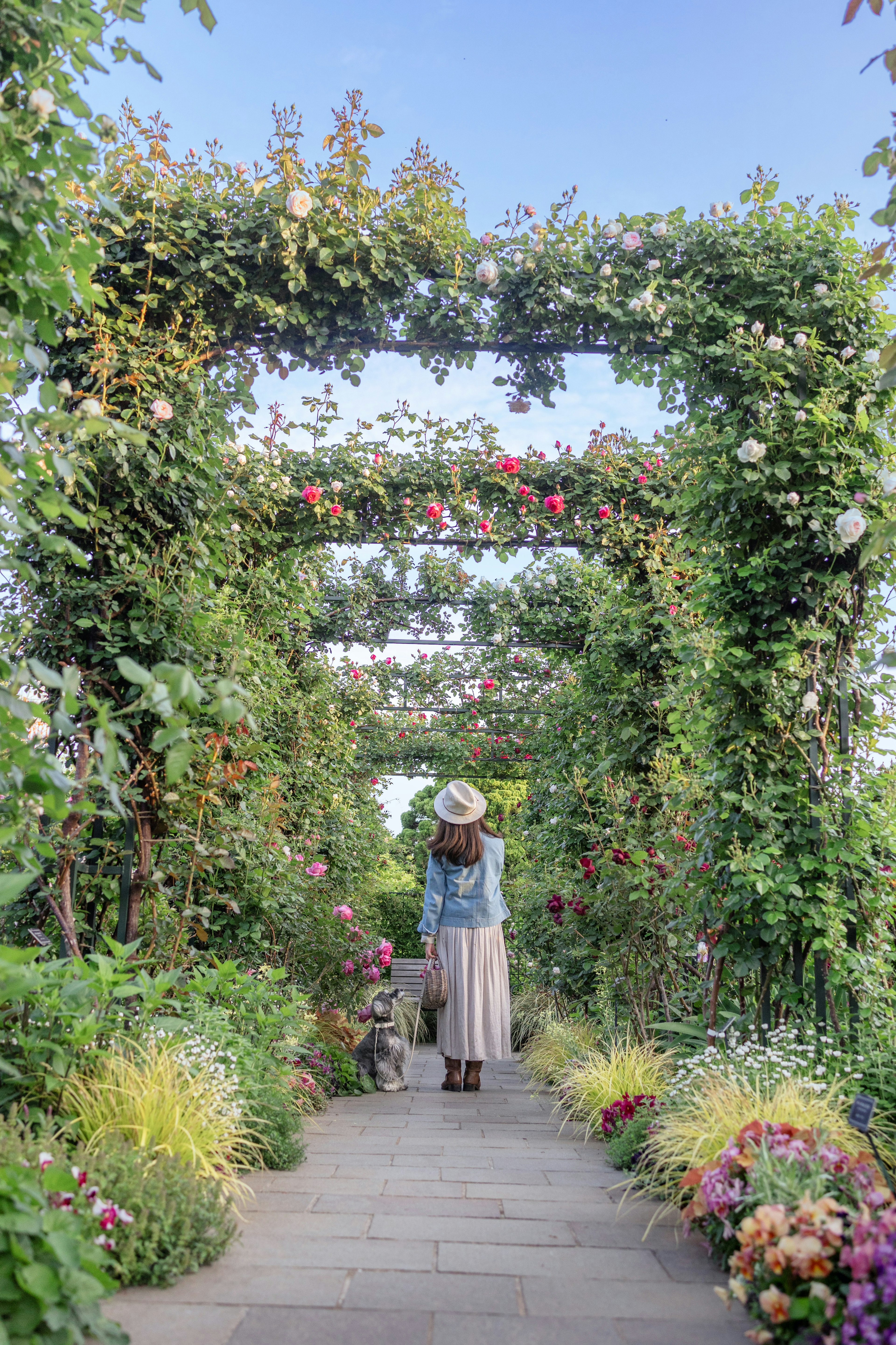 Mujer de pie bajo un arco de jardín exuberante rodeada de plantas y flores