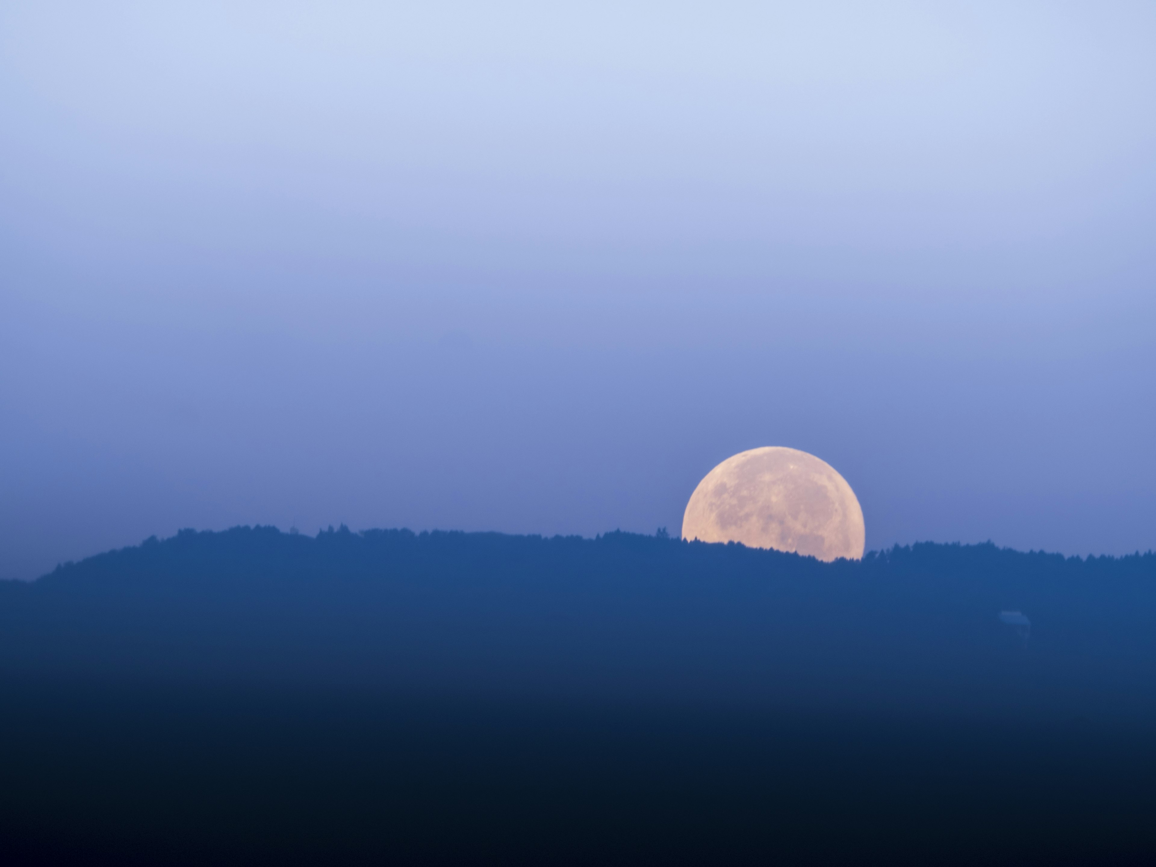 Large full moon rising above mountain silhouette under blue sky