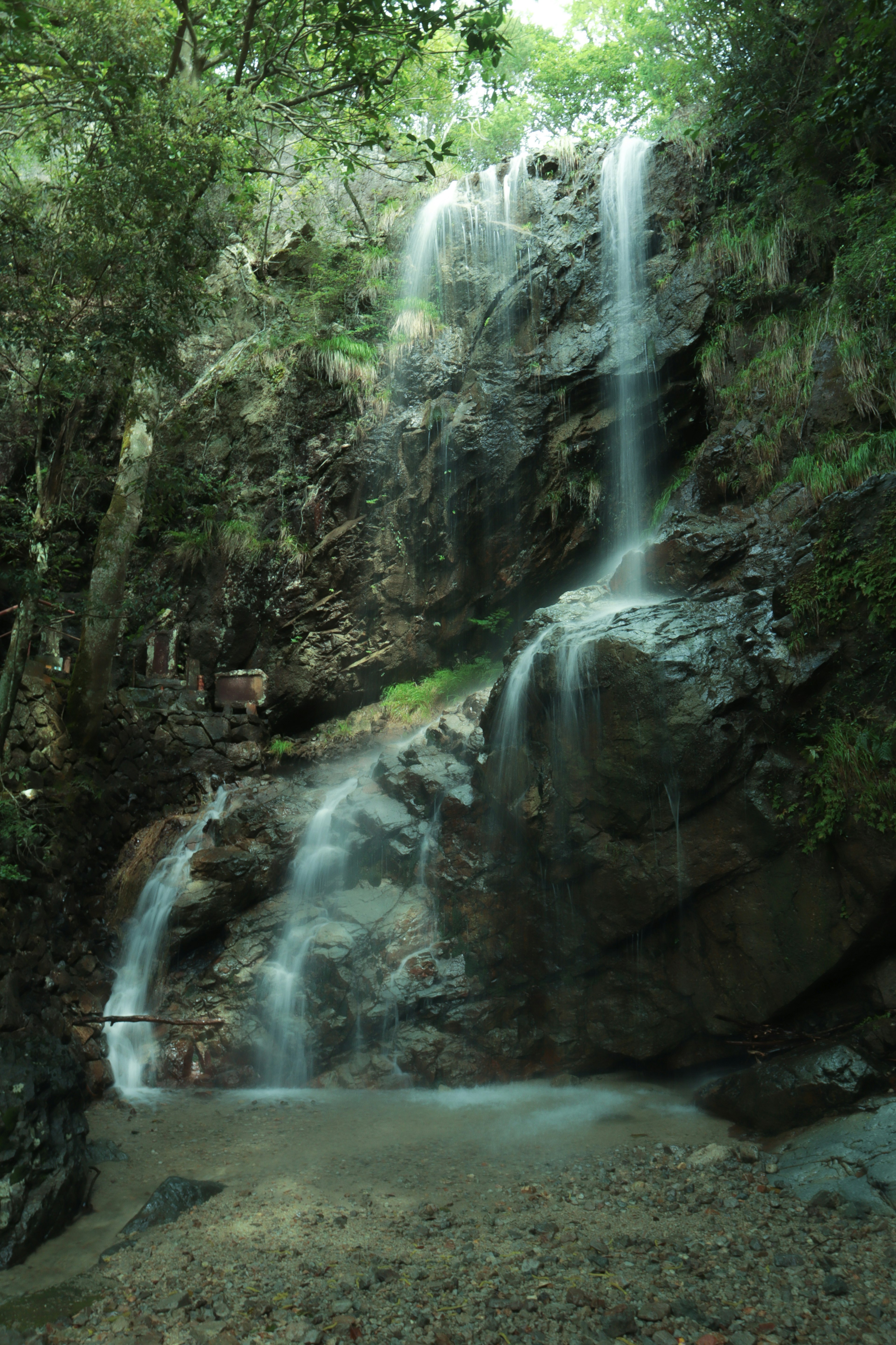 Sebuah air terjun mengalir turun dari medan berbatu dikelilingi oleh kehijauan yang rimbun