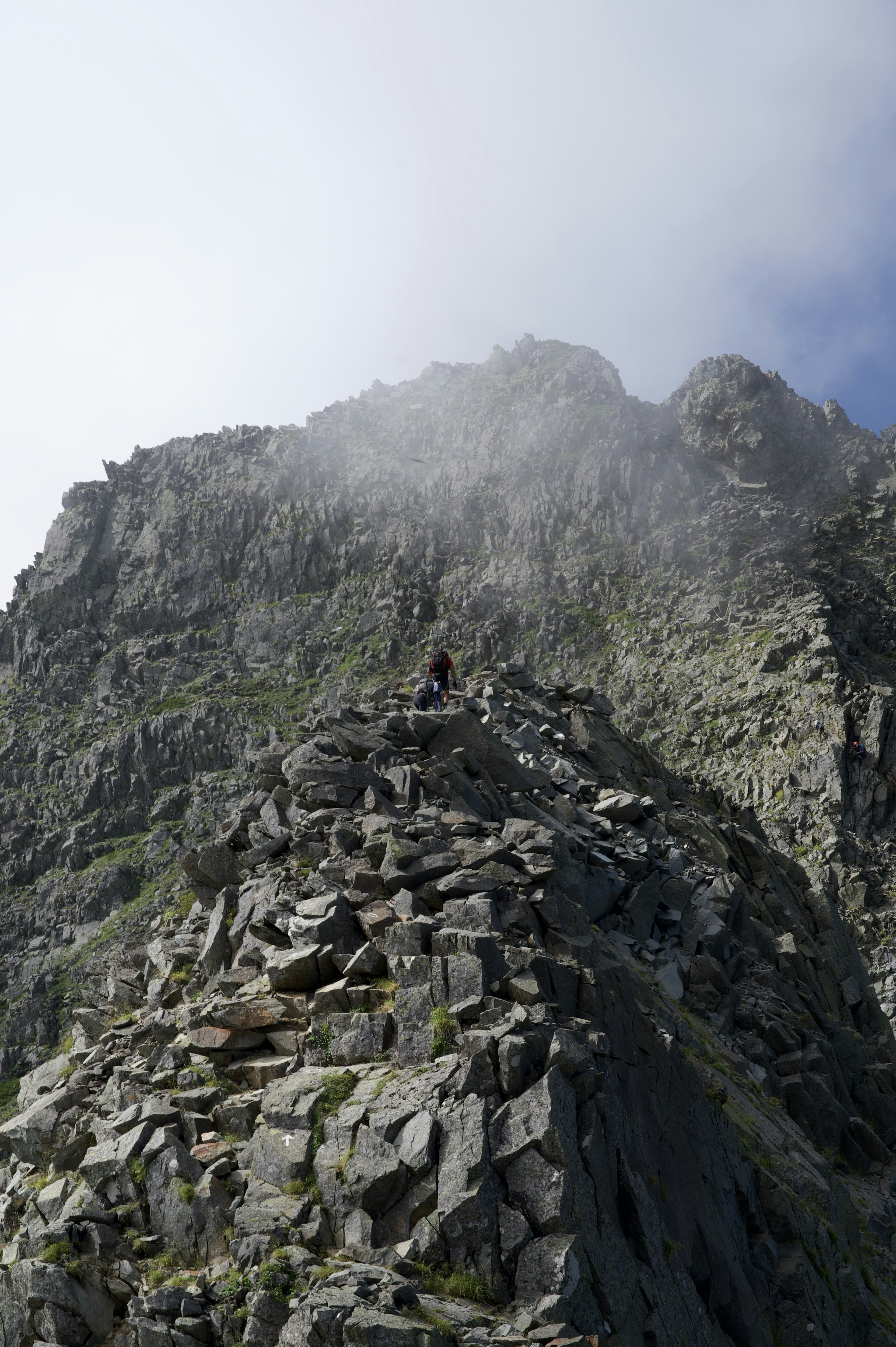 Escursionista che sale verso una cima montuosa rocciosa con nebbia e nuvole