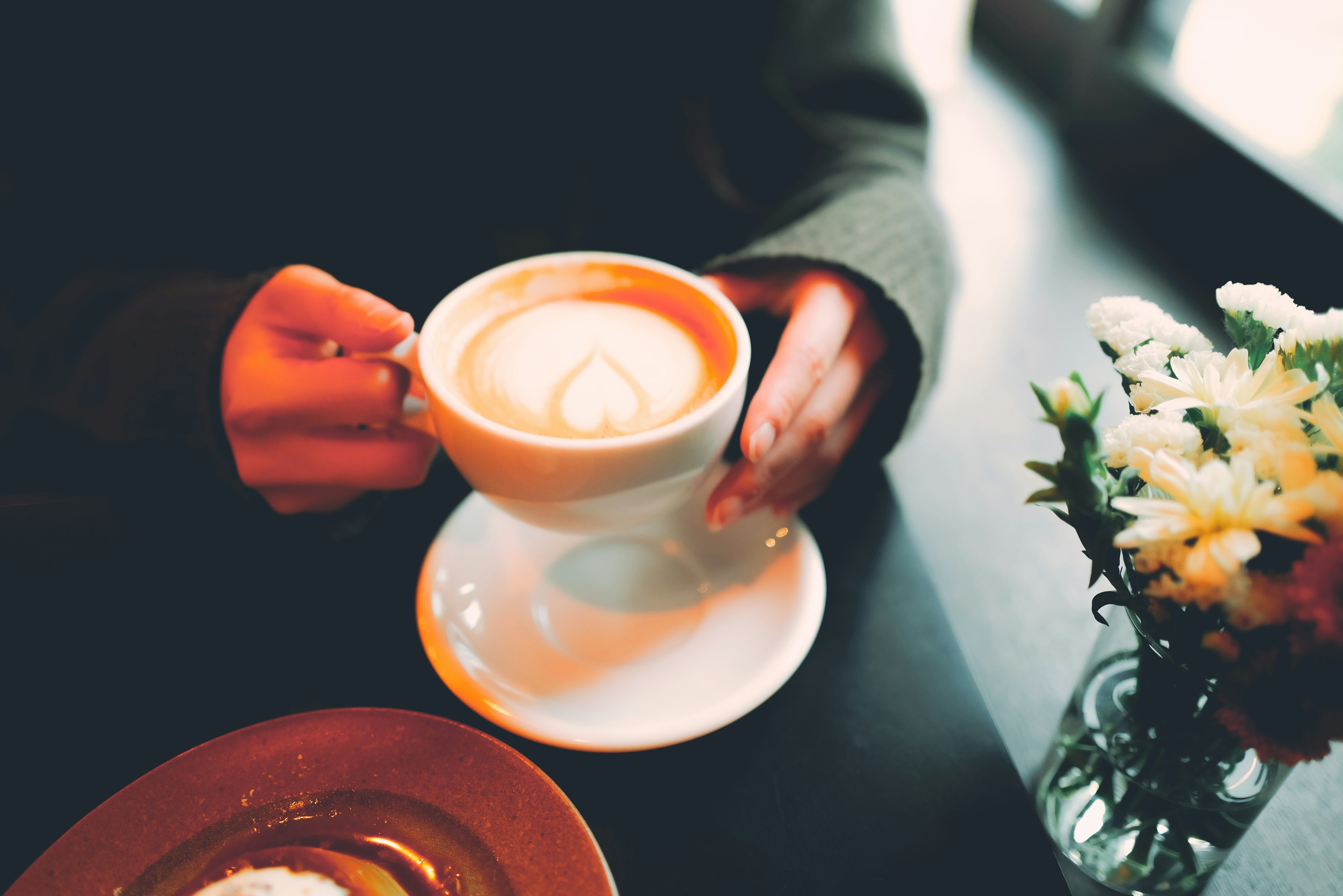 Hand hält eine Tasse mit Latte-Art und Blumen in einer Vase