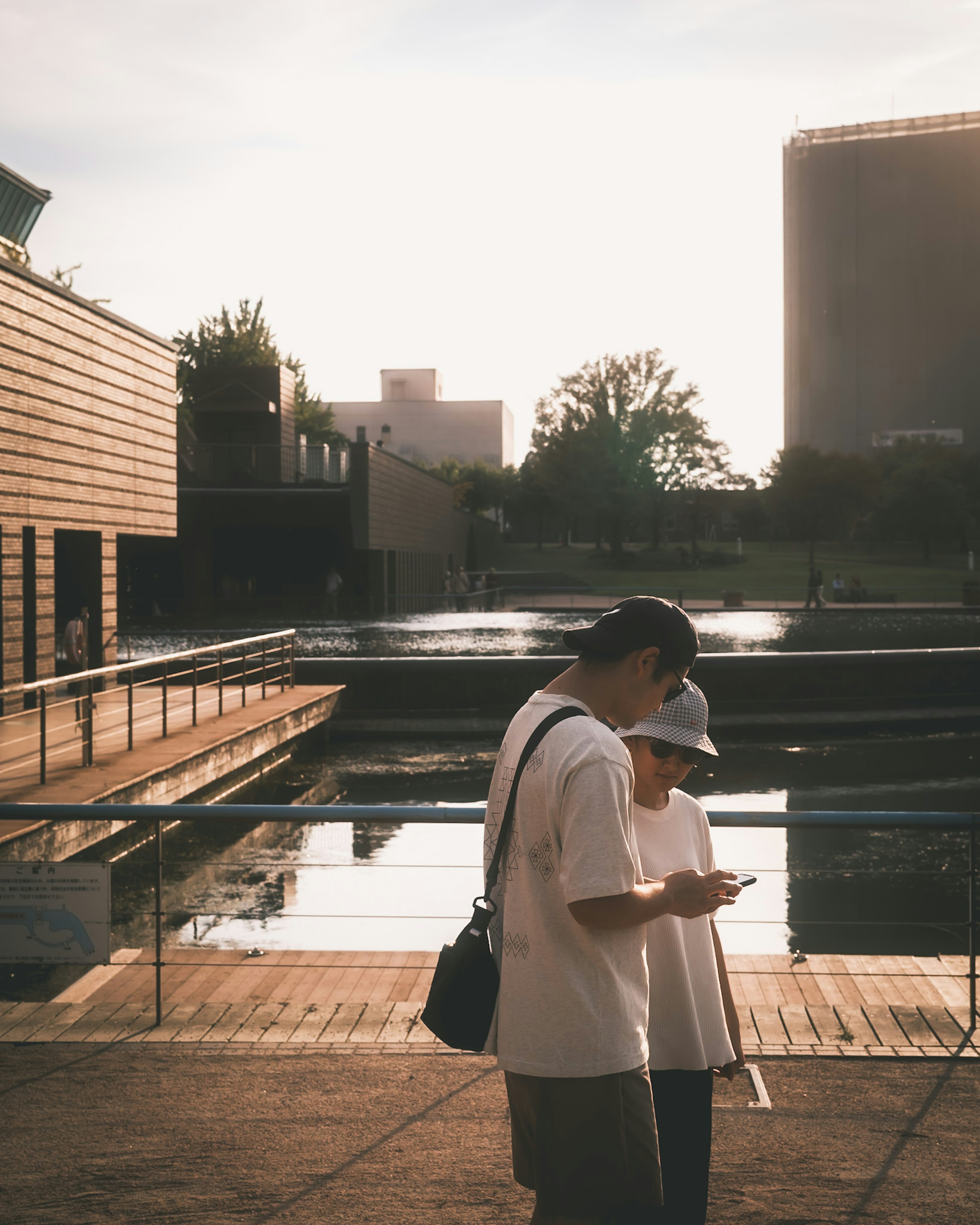 夕暮れ時の公園でスマートフォンを使う二人の人物と水面に映る建物