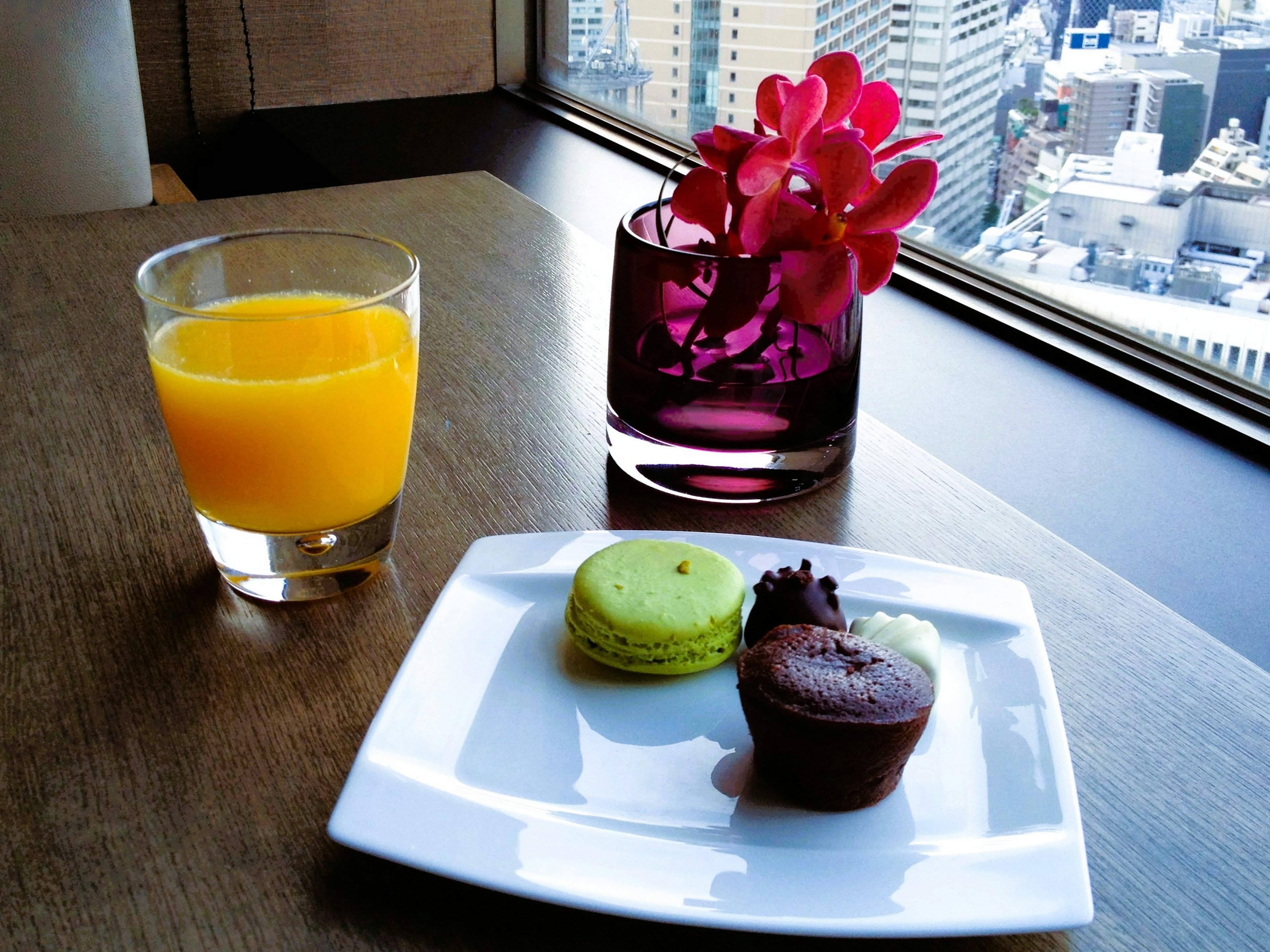 Un piatto con un macaron verde e un cupcake al cioccolato con un bicchiere di succo d'arancia e fiori viola vicino alla finestra