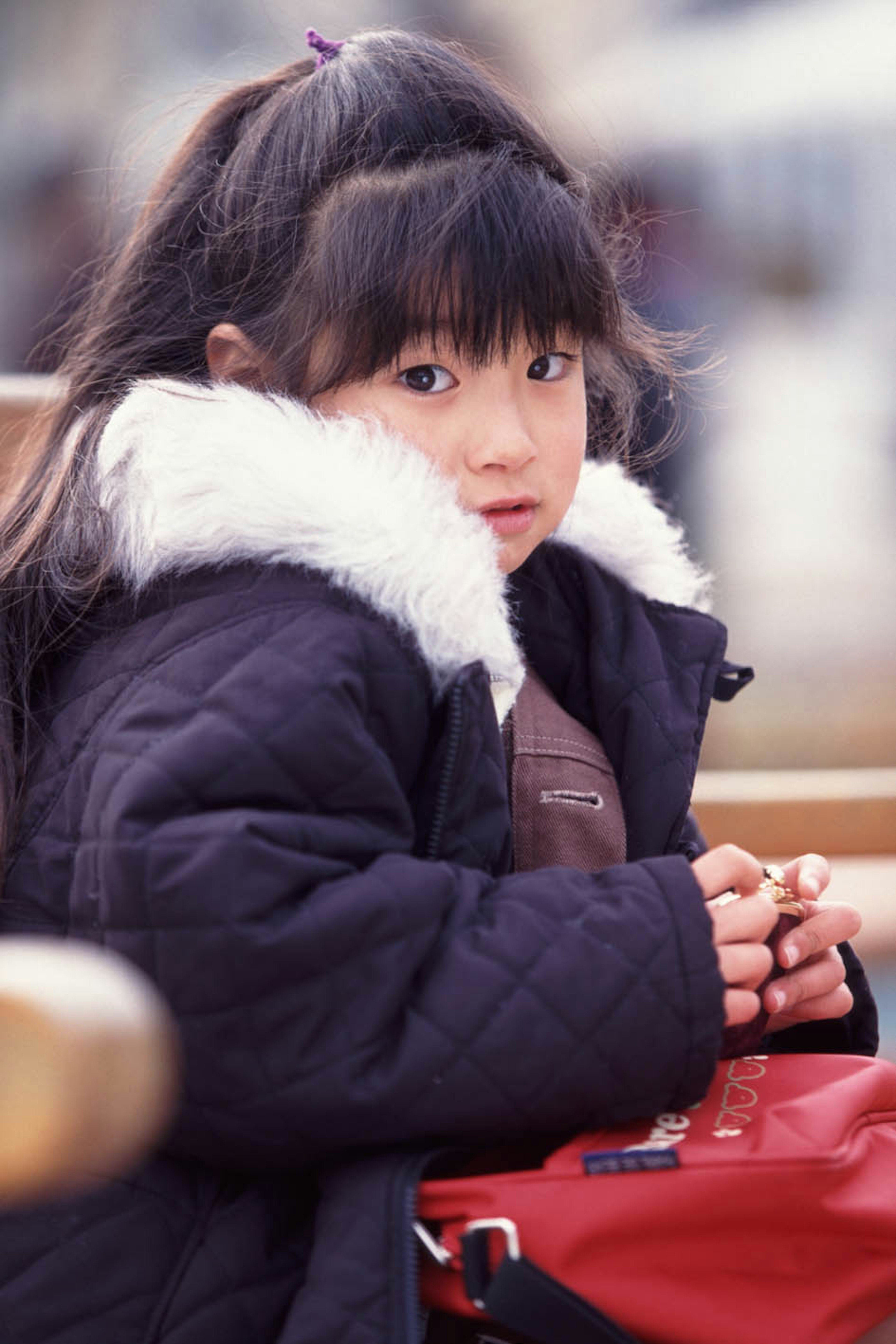 Una niña con abrigo negro está sentada con cabello largo y negro y un cuello blanco esponjoso