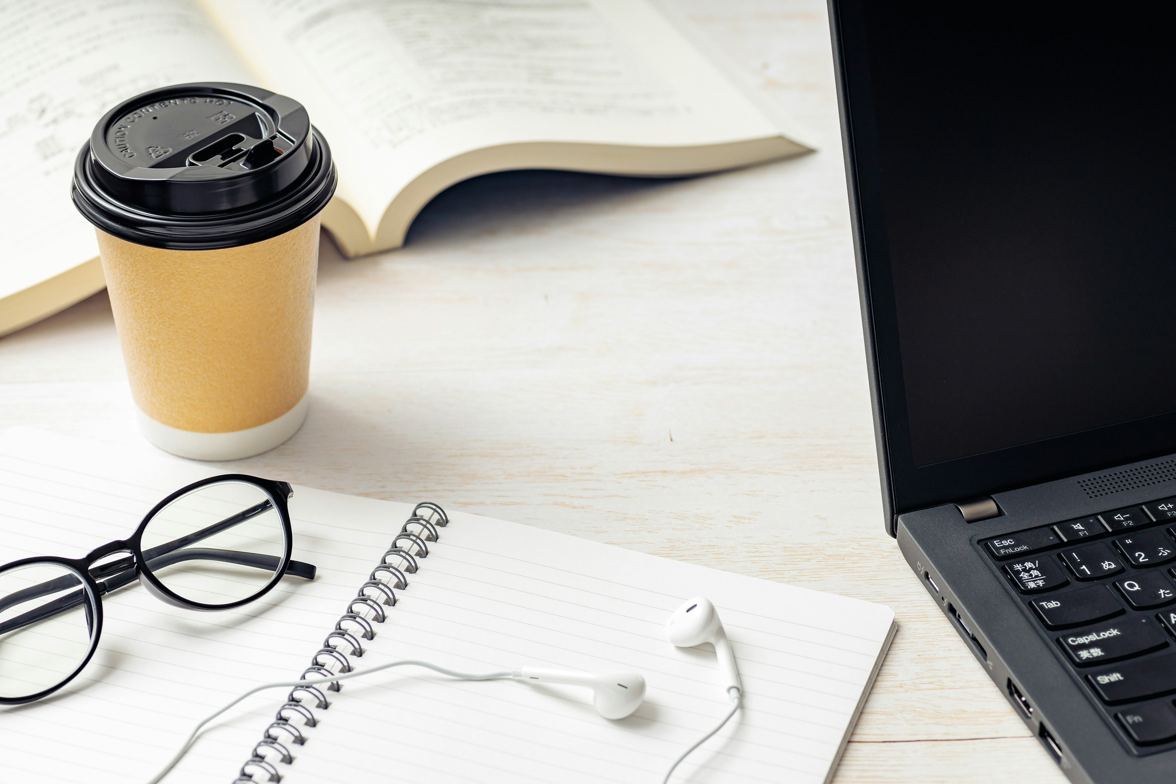 Coffee cup, laptop, notebook, glasses, and earbuds on a desk