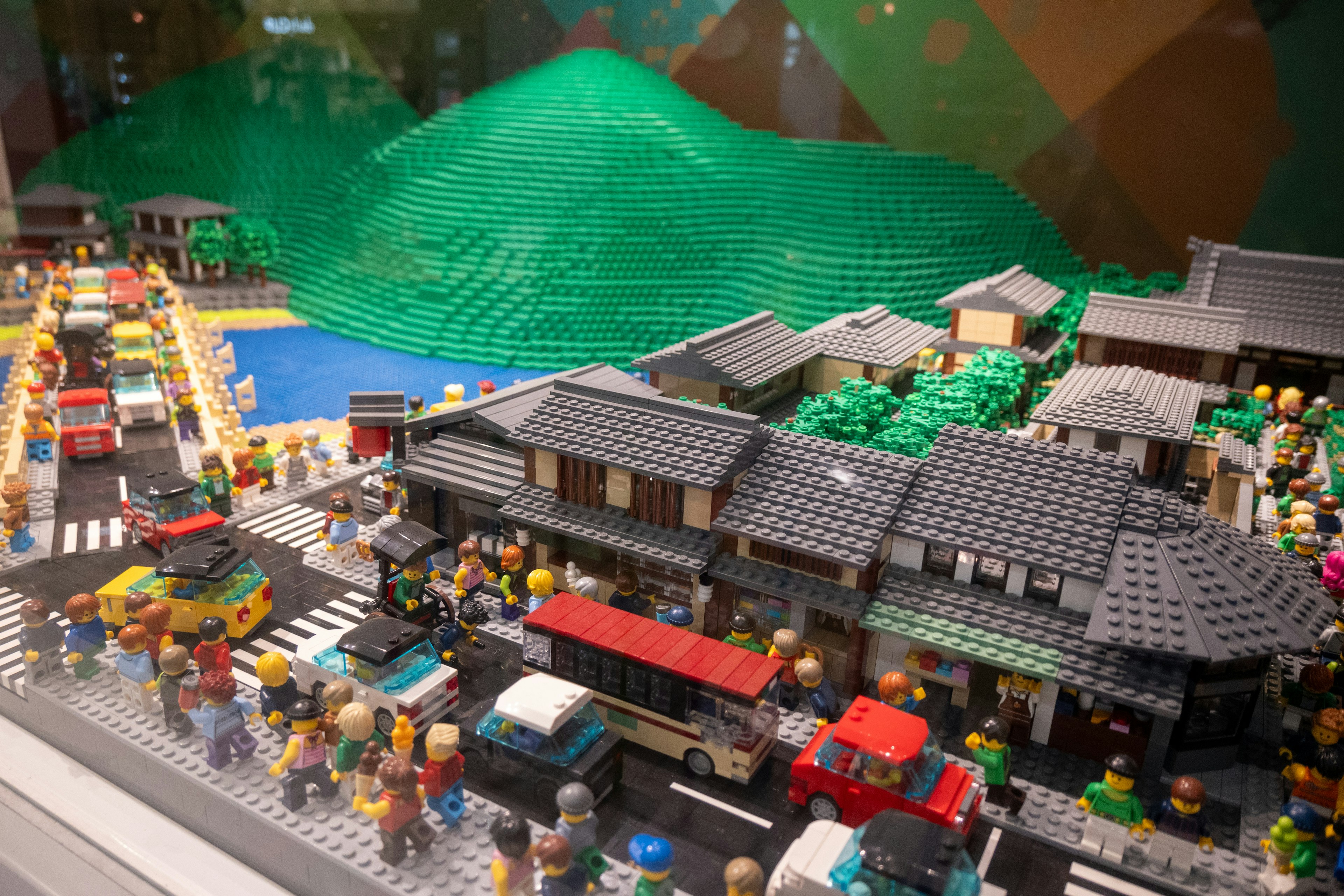 Lego cityscape model featuring buildings vehicles and people with a green mountain backdrop