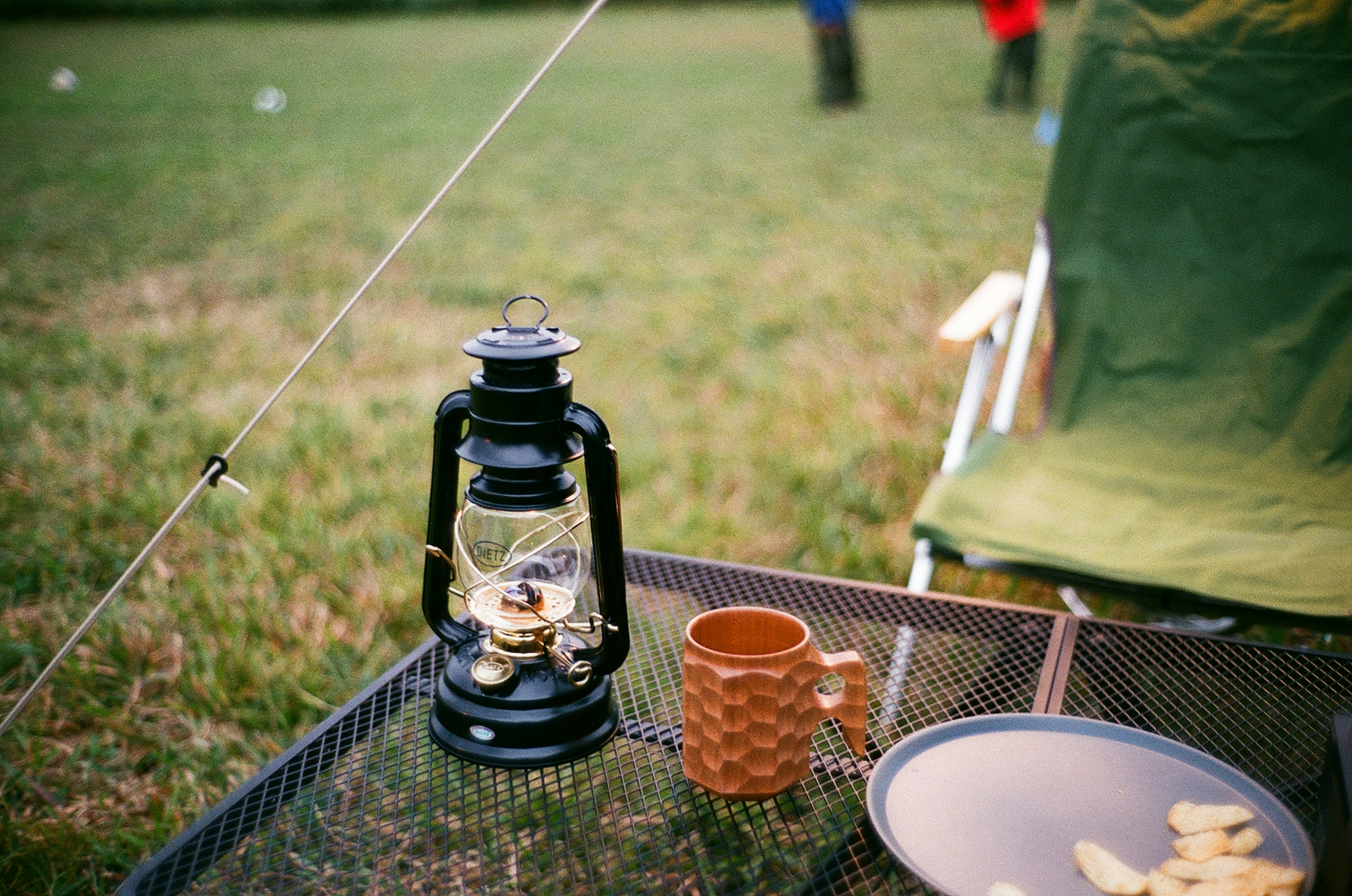 Linterna negra y taza naranja en una mesa de camping con fondo de hierba