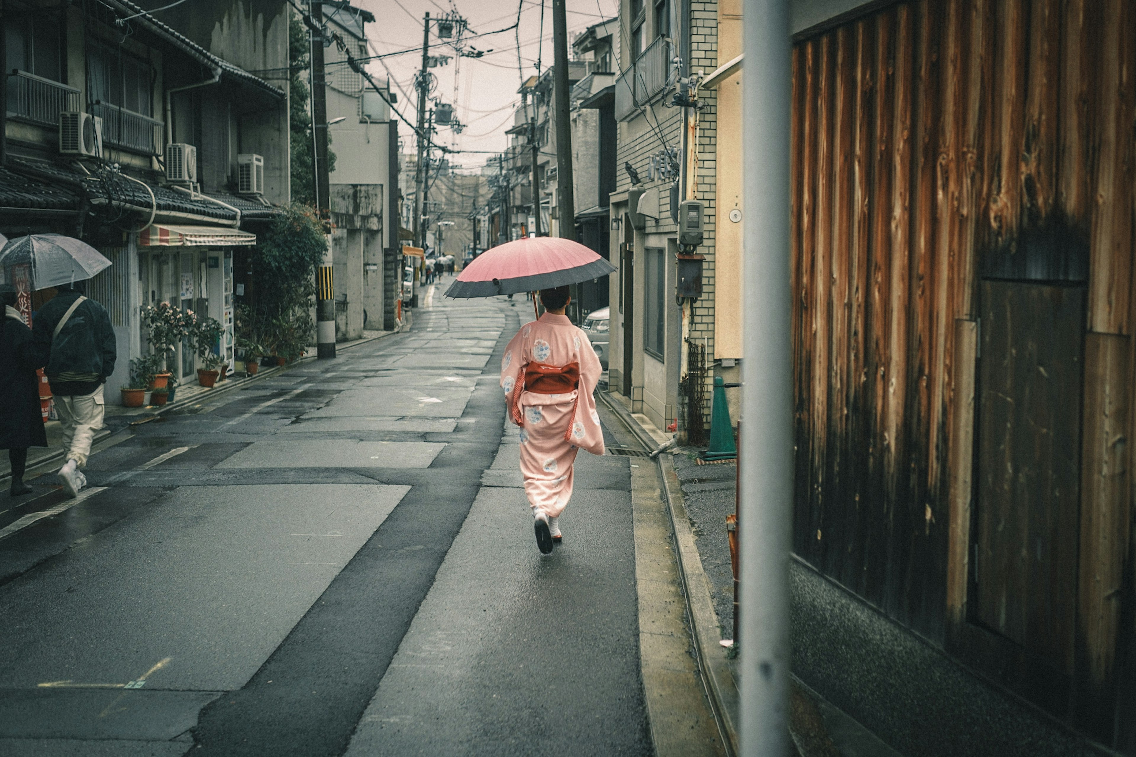 Wanita berpakaian kimono berjalan dengan payung merah muda di jalan yang hujan