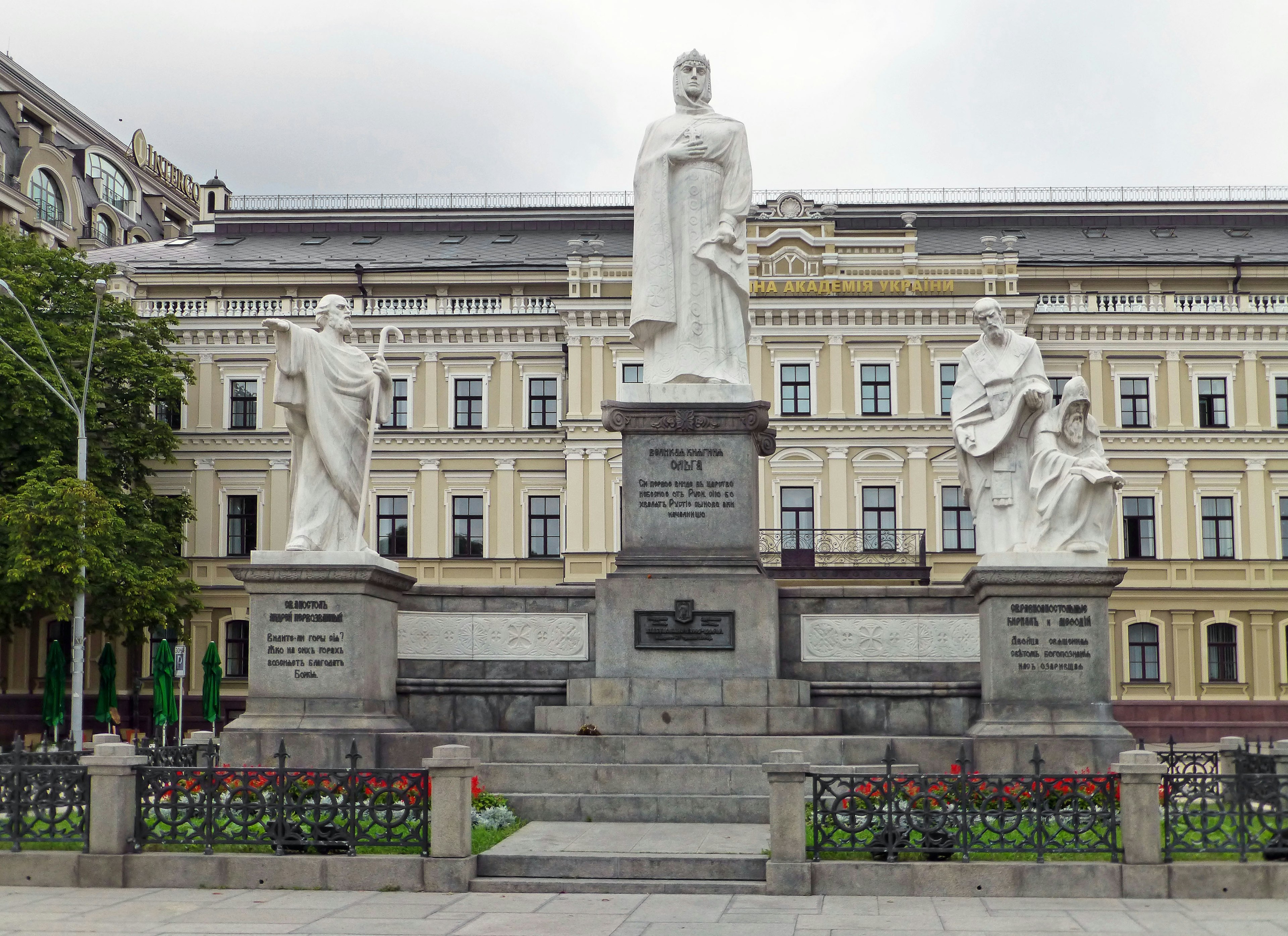 Monument with white statues surrounded by greenery and historic building in the background