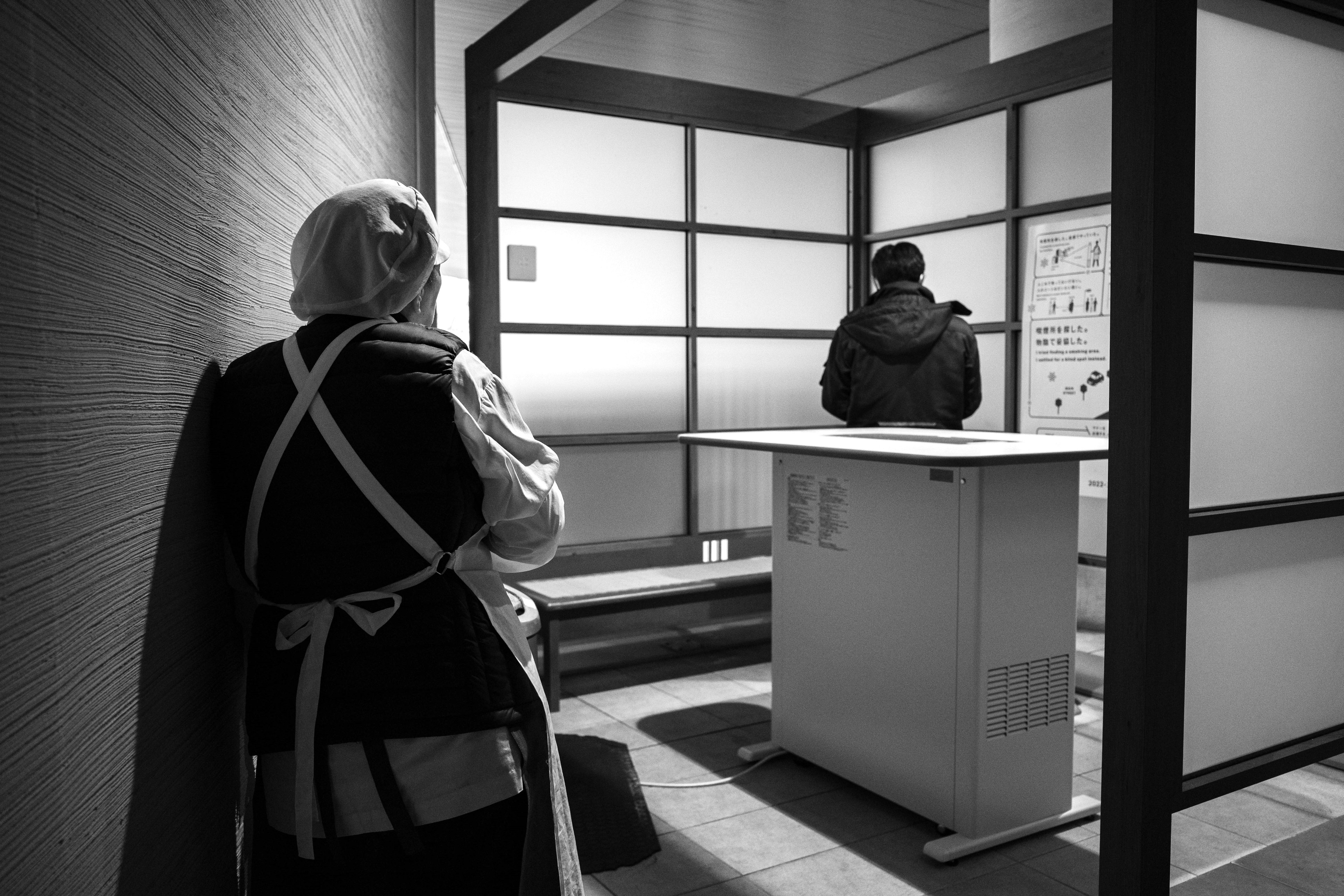 Scène en noir et blanc avec une personne en tenue traditionnelle observant une autre personne à une table blanche