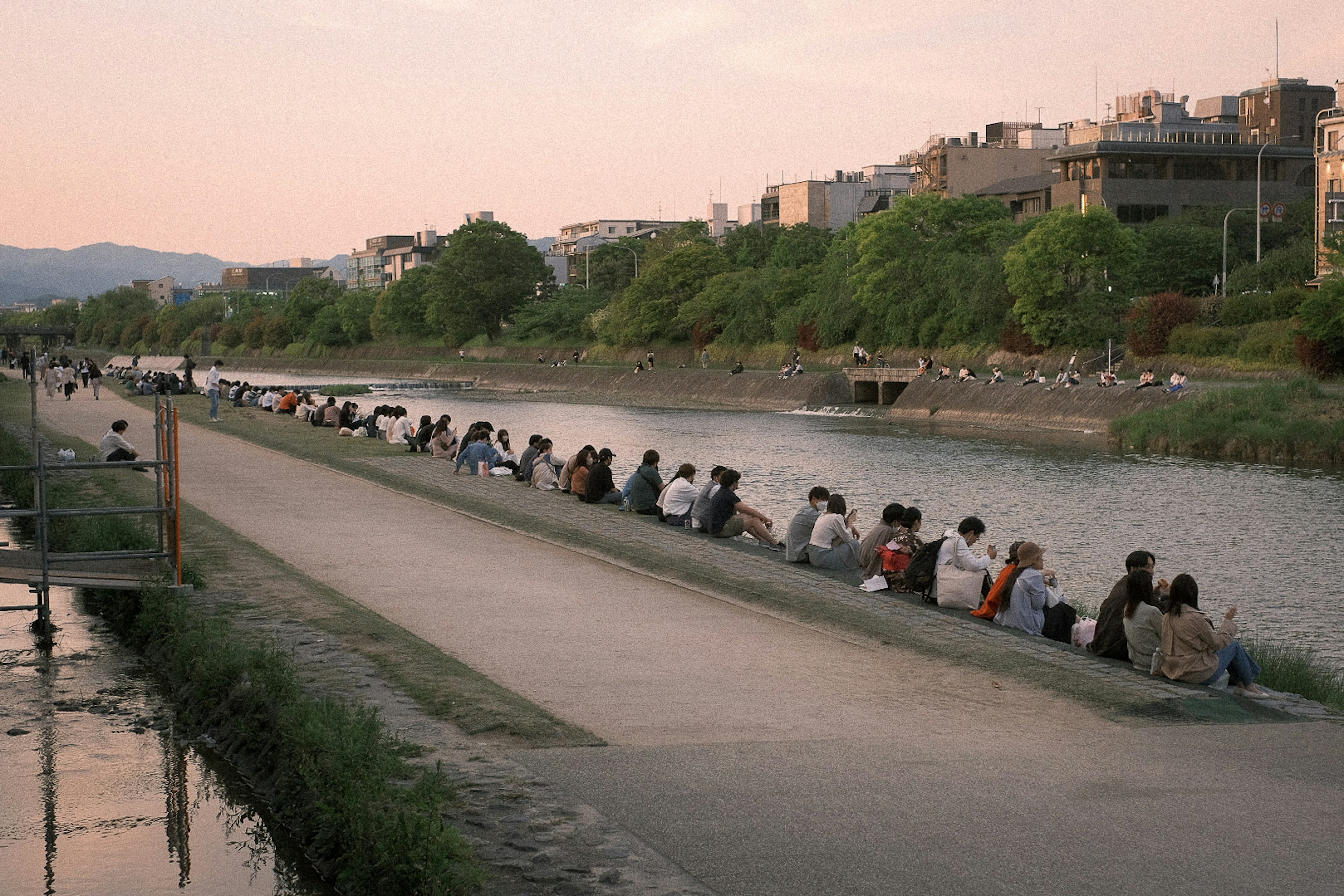 Row of people sitting by the river at sunset with lush greenery and buildings in the background