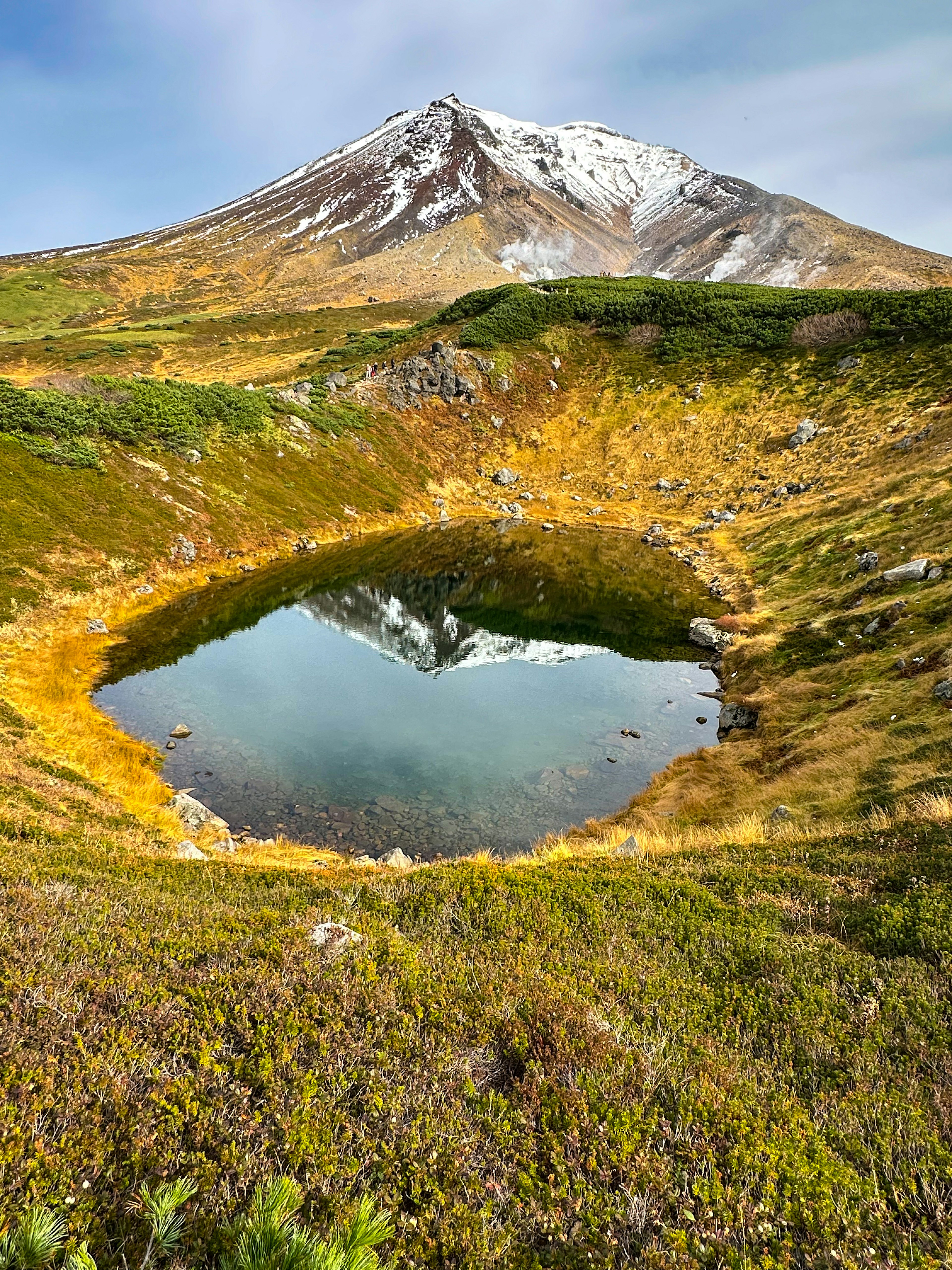 Ein kleiner Teich umgeben von grünen und gelben Wiesen mit einem schneebedeckten Berg im Hintergrund