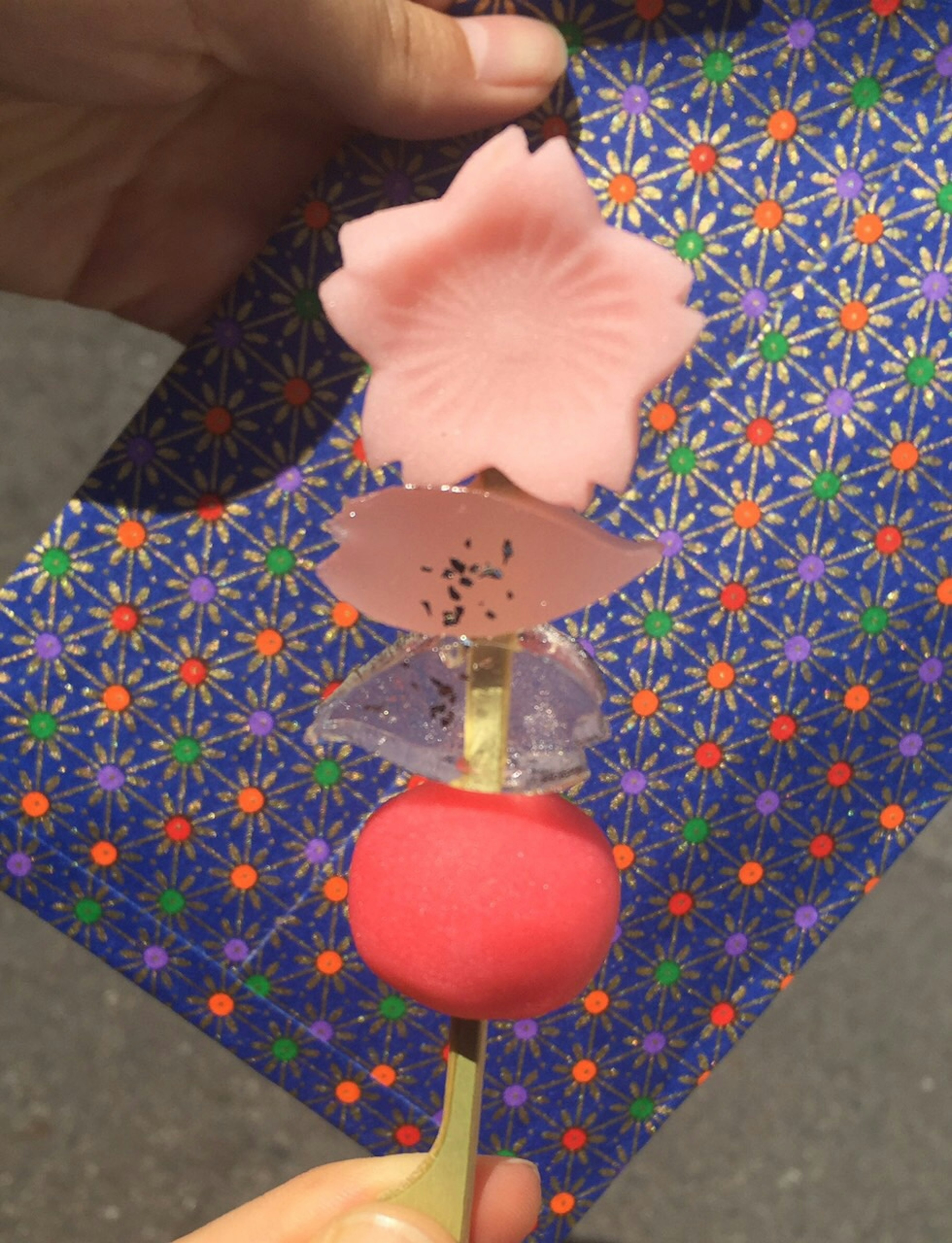 A skewer of Japanese sweets featuring a cherry blossom shape and colorful dango with a vibrant patterned bag