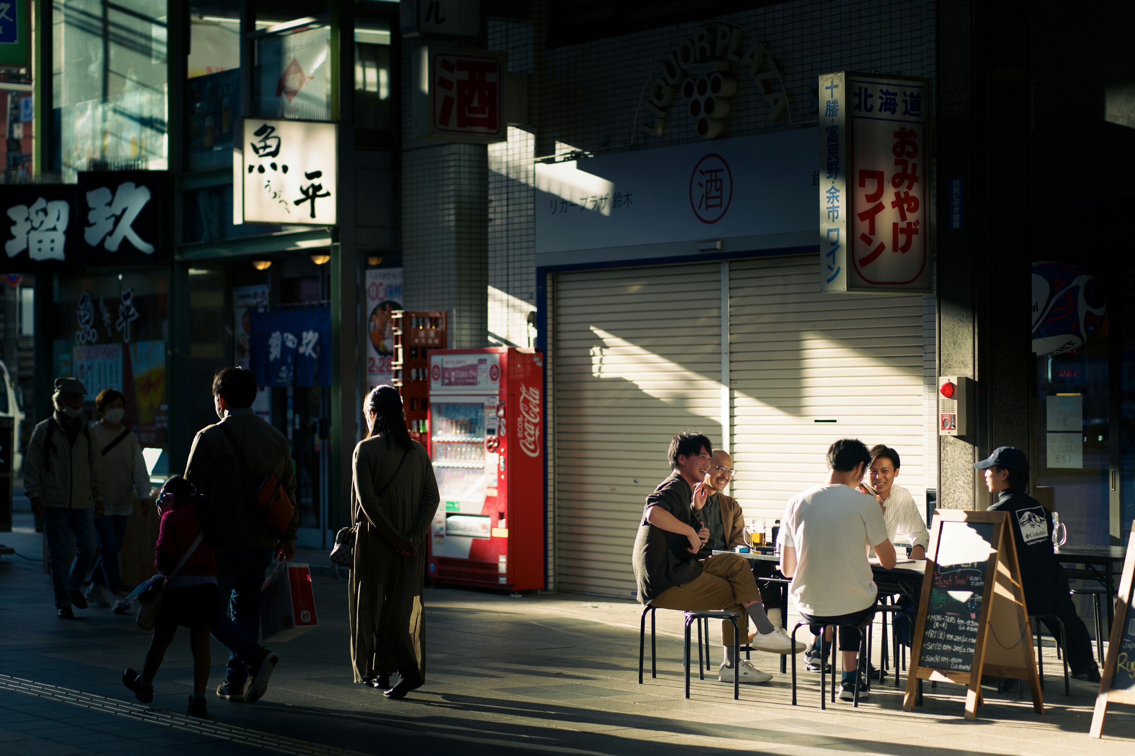 Escena de personas disfrutando de un café al sol con peatones en una calle concurrida