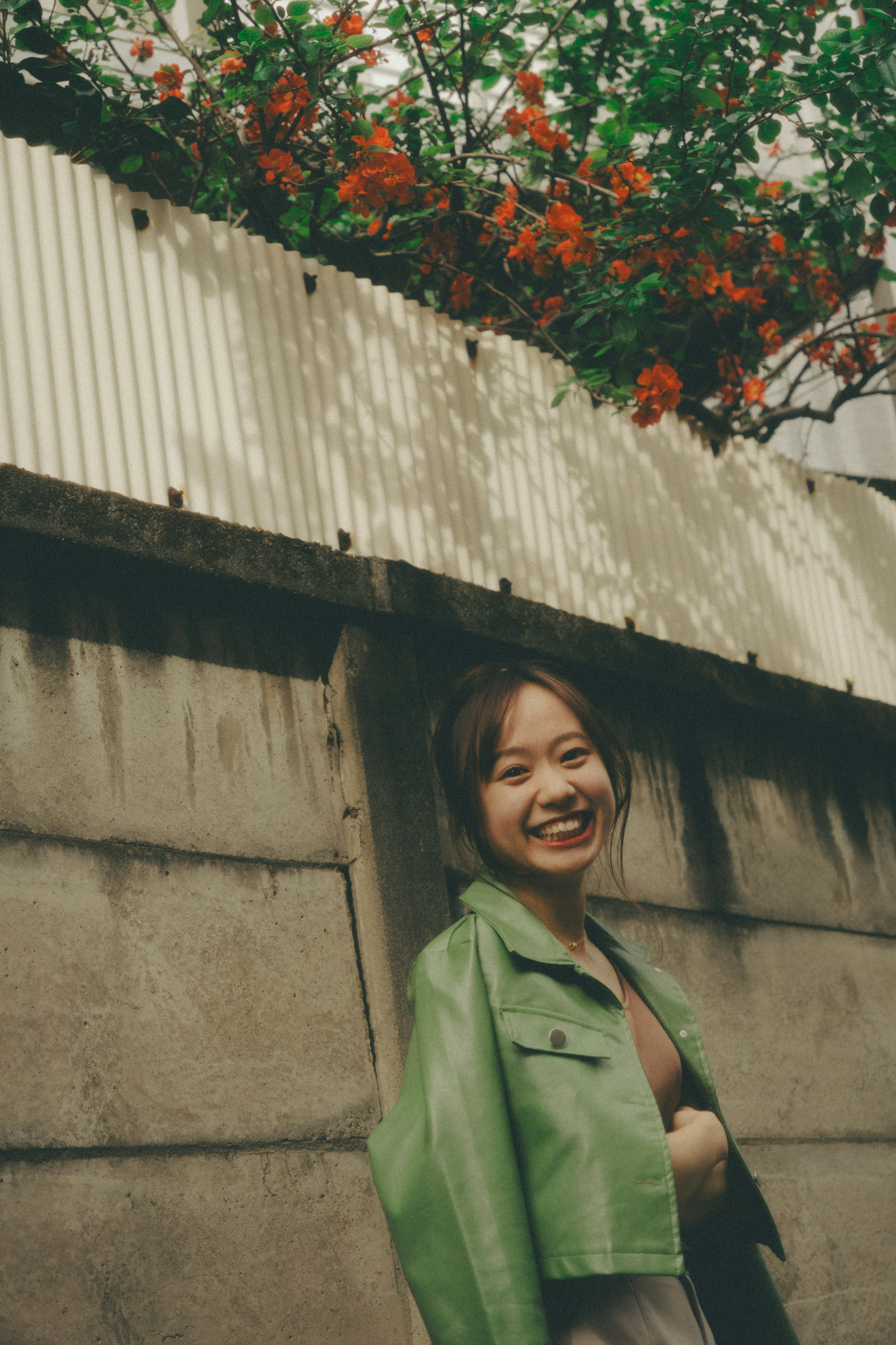 A smiling woman in a green jacket stands by a wall with blooming flowers in the background