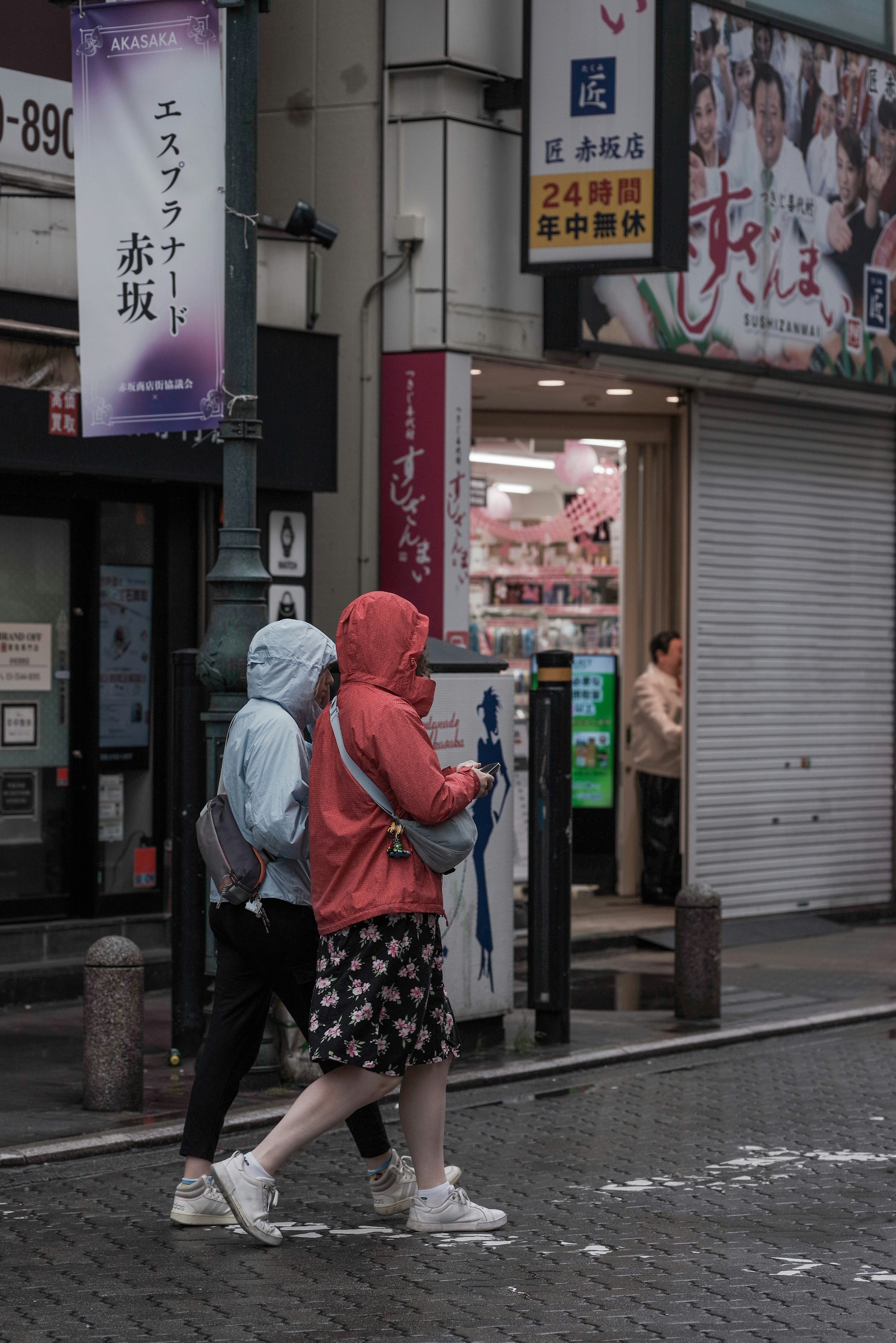 Deux femmes marchant sous la pluie L'une porte un imperméable bleu et l'autre une veste à capuche rouge Des enseignes de magasin sont visibles en arrière-plan