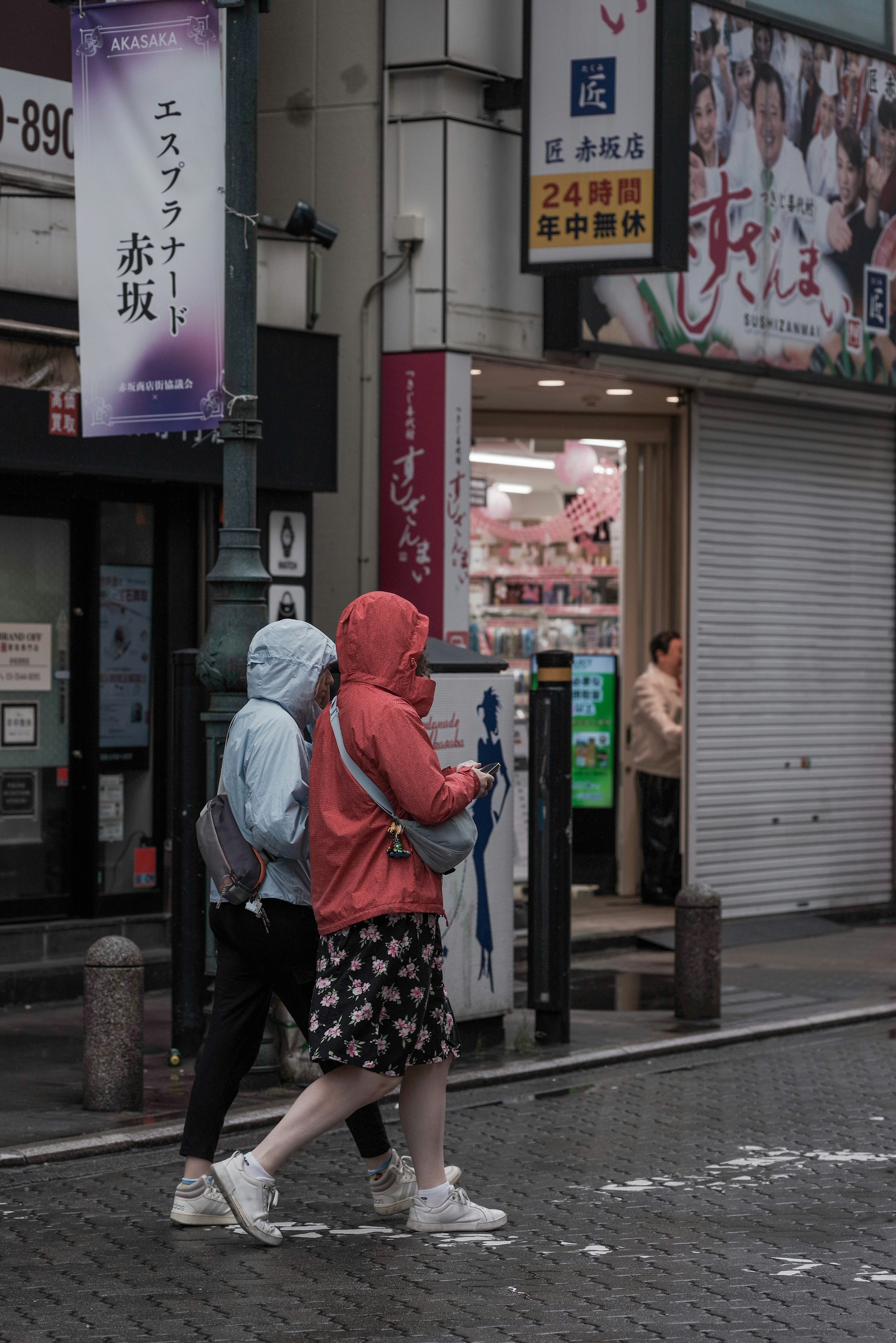 Deux femmes marchant sous la pluie L'une porte un imperméable bleu et l'autre une veste à capuche rouge Des enseignes de magasin sont visibles en arrière-plan