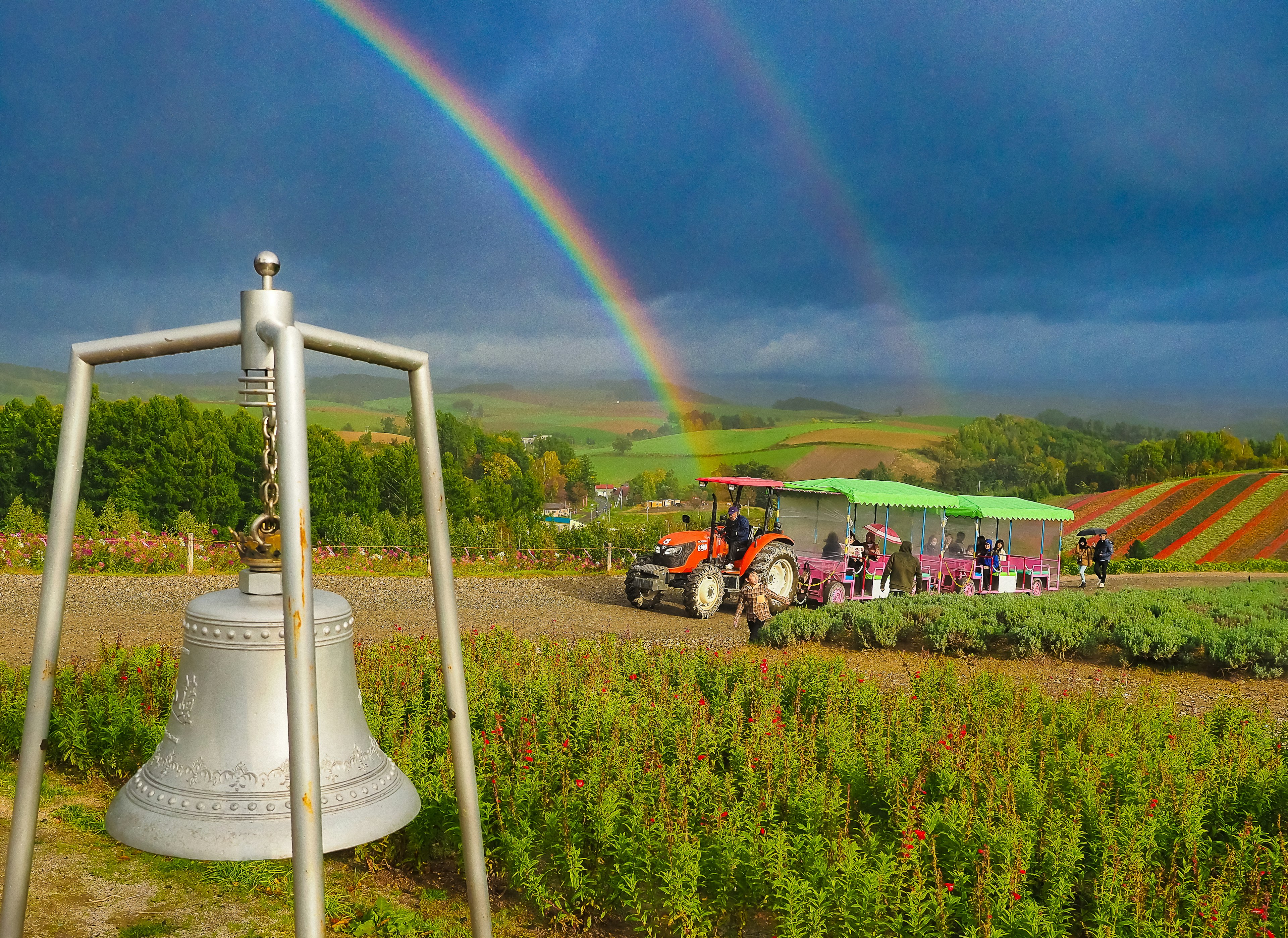 Touristen besuchen einen Bauernhof unter einem Regenbogen mit einem Traktor bunten Blumenfeldern und einer großen Glocke