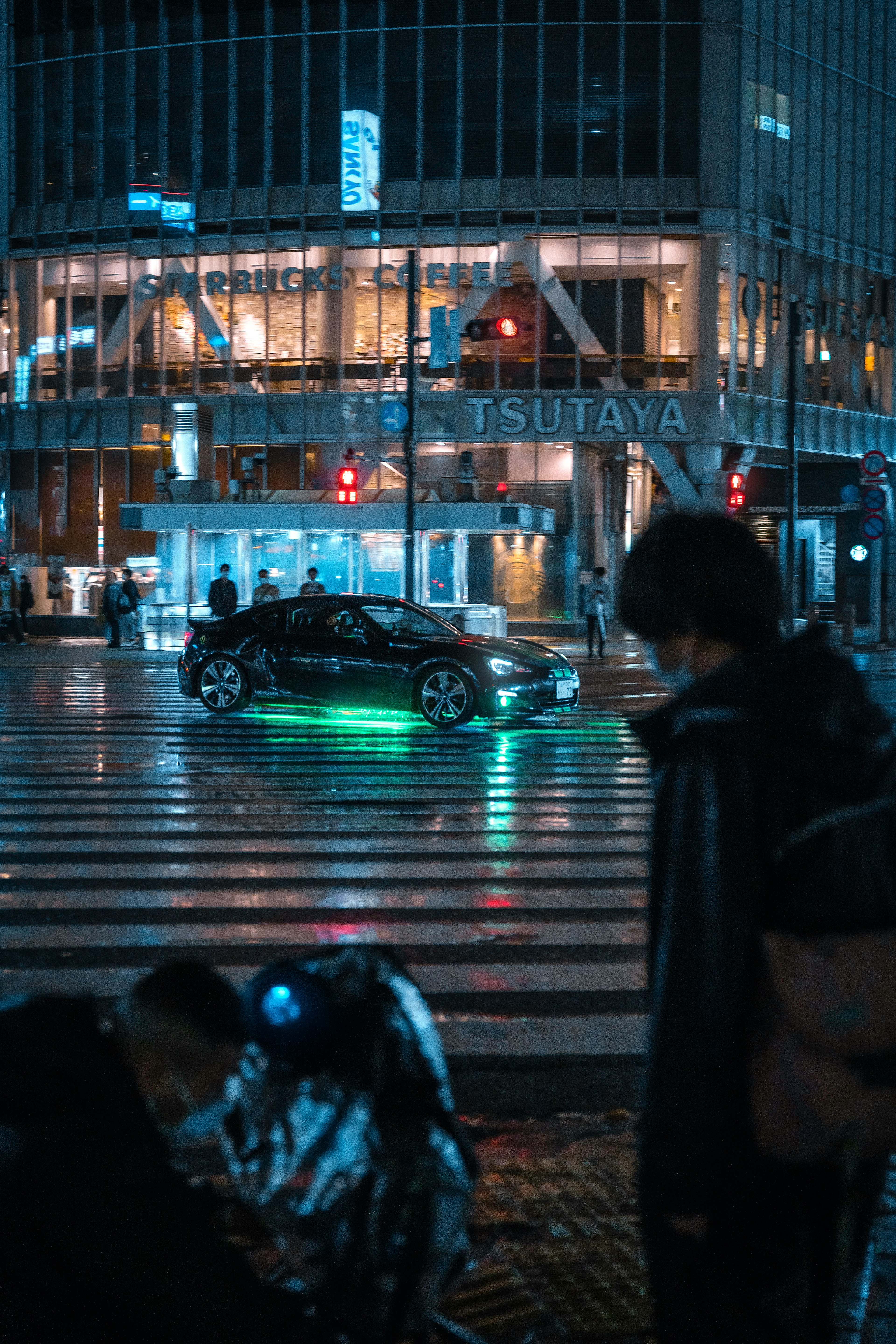 People at a nighttime crosswalk with a car featuring green lights