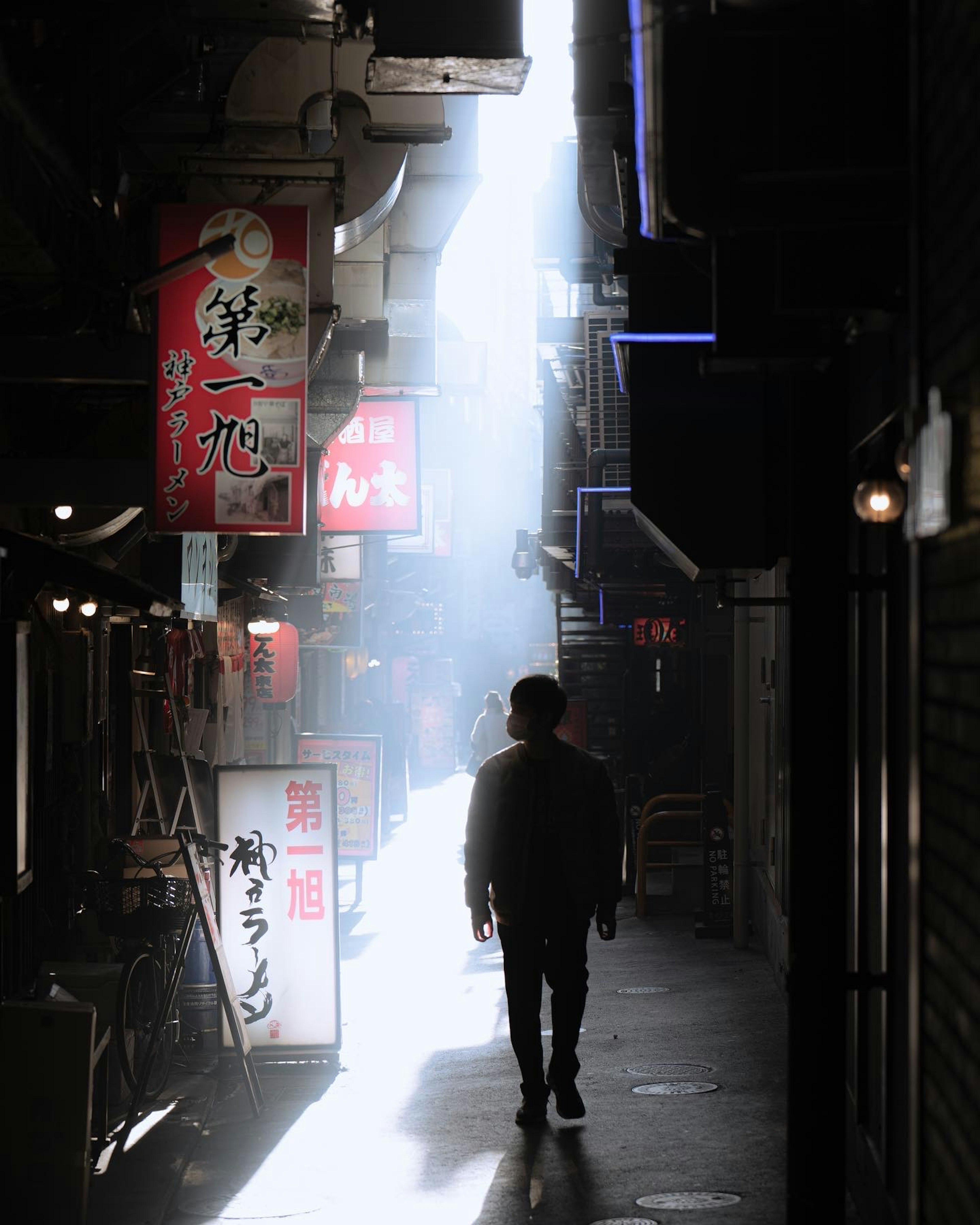 A silhouette of a person walking in a dark alley illuminated by a beam of light and red signs