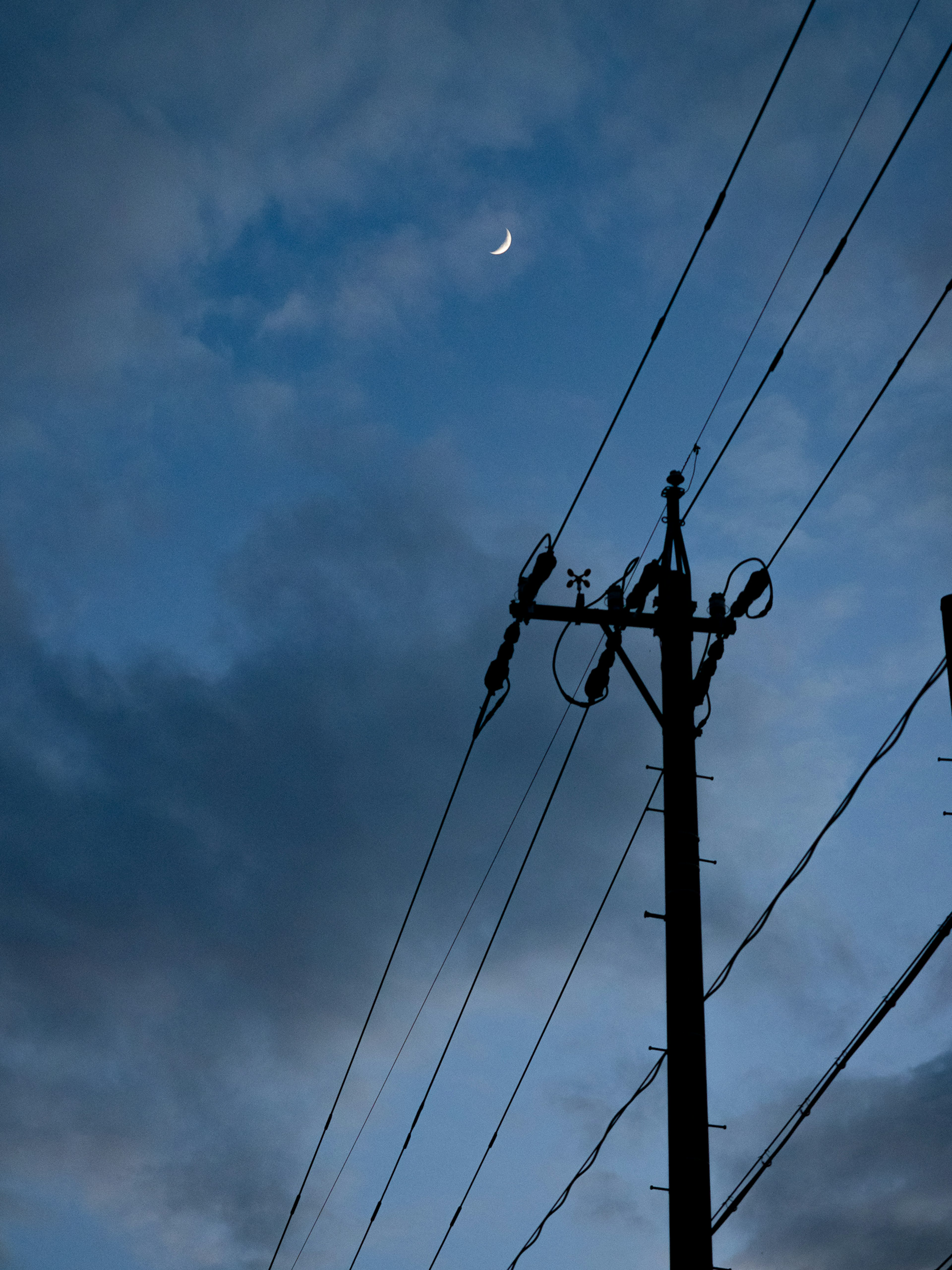 Silhouette eines Strommastes mit Stromleitungen vor einem Dämmerungshimmel mit einem zunehmenden Mond