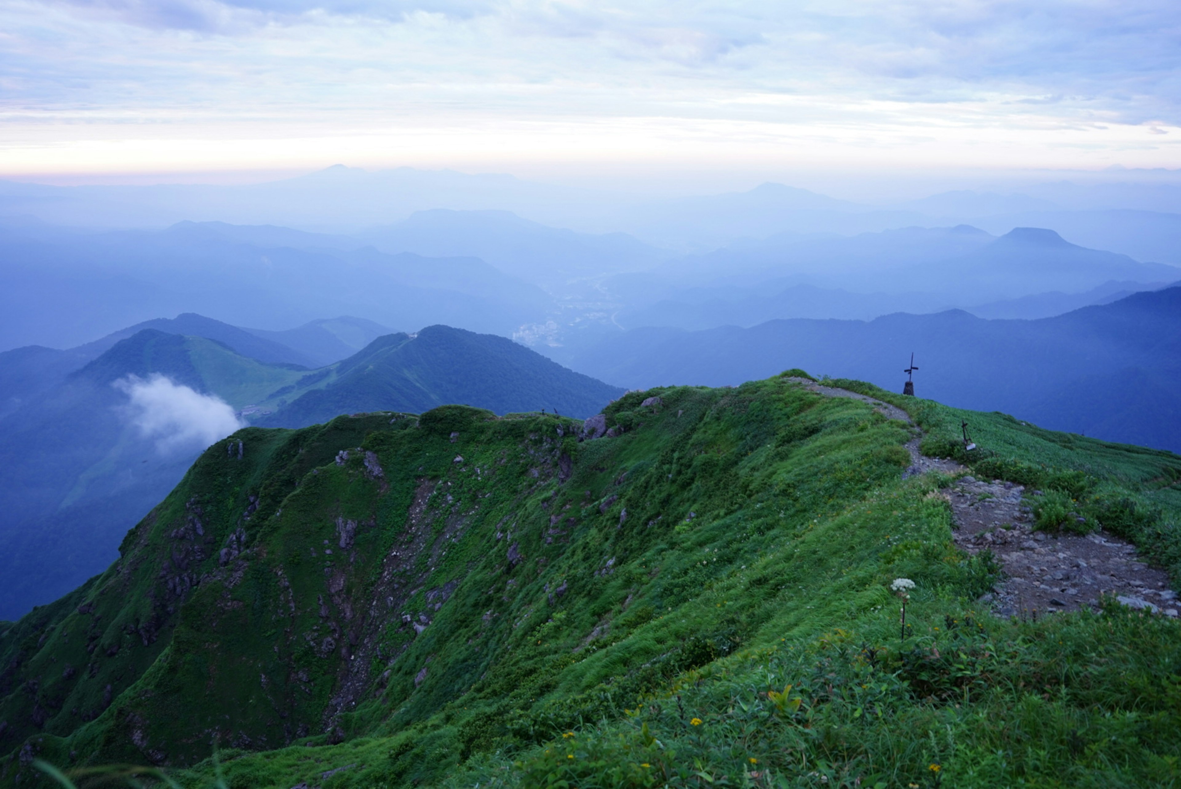 Puncak gunung dengan rumput hijau subur dan pegunungan jauh di bawah langit biru