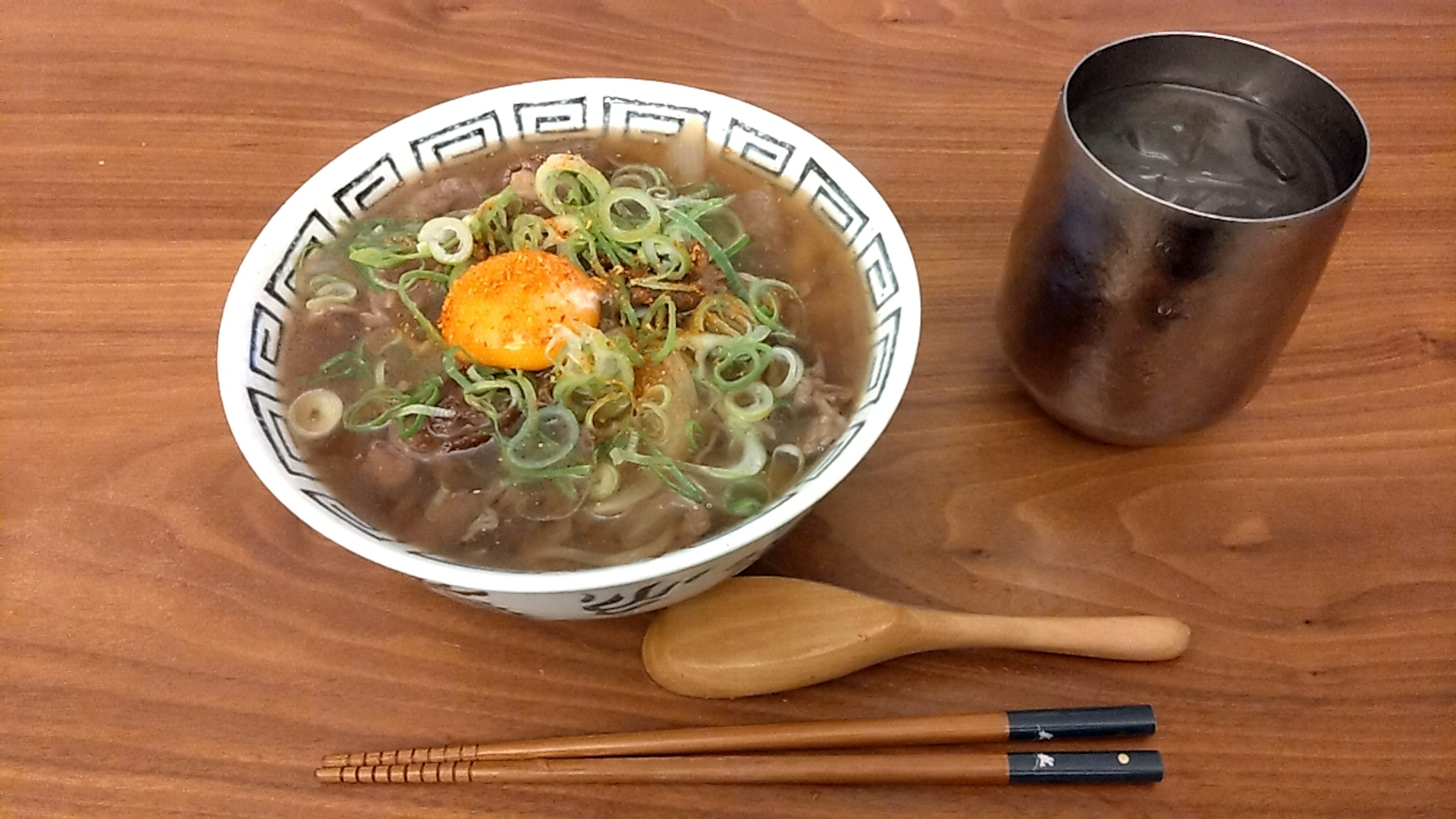 Mangkok ramen dengan bawang hijau dan telur mentah di atasnya disertai sendok kayu dan sumpit di atas meja kayu