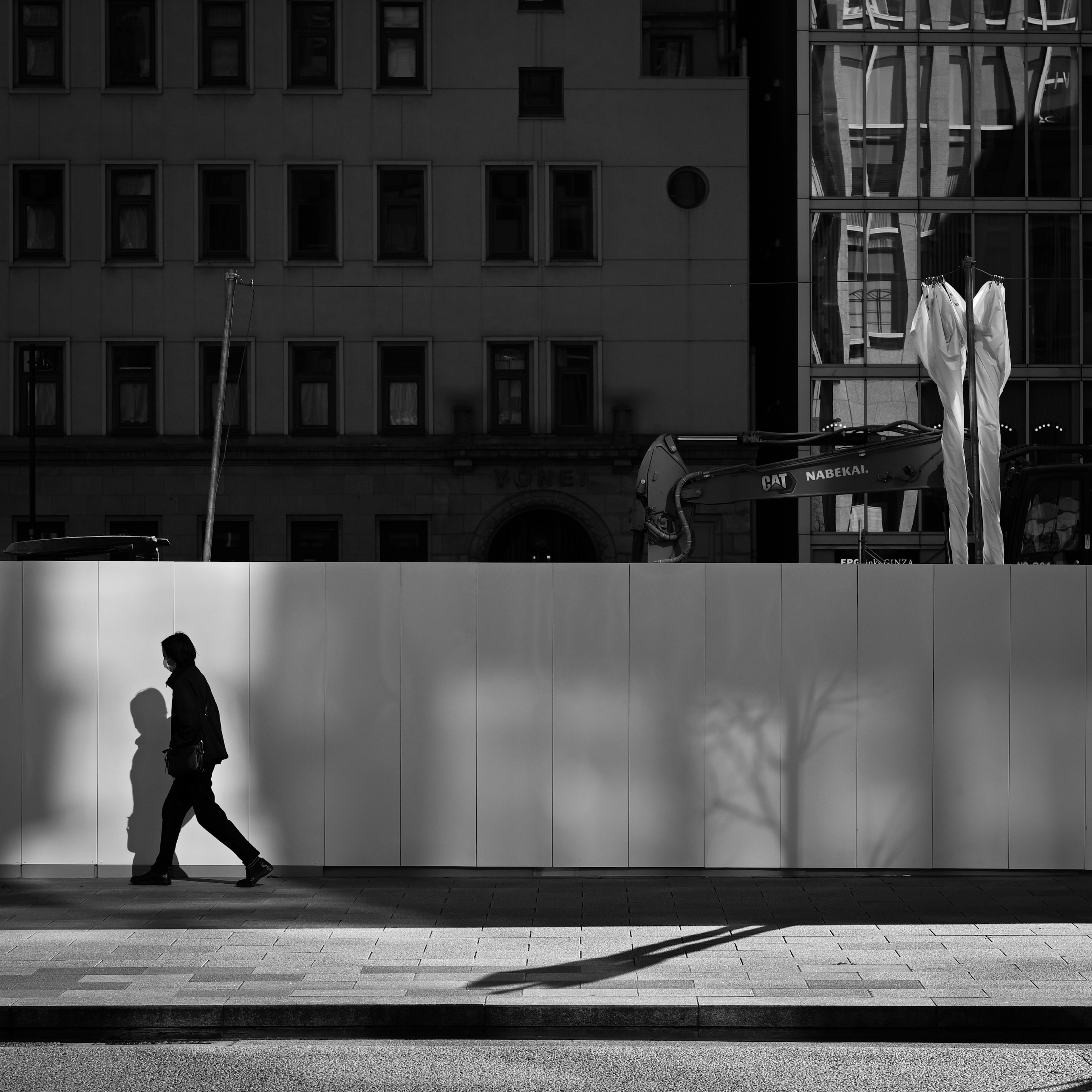 A black and white urban scene featuring a person walking with shadows and striking buildings and sculptures in the background