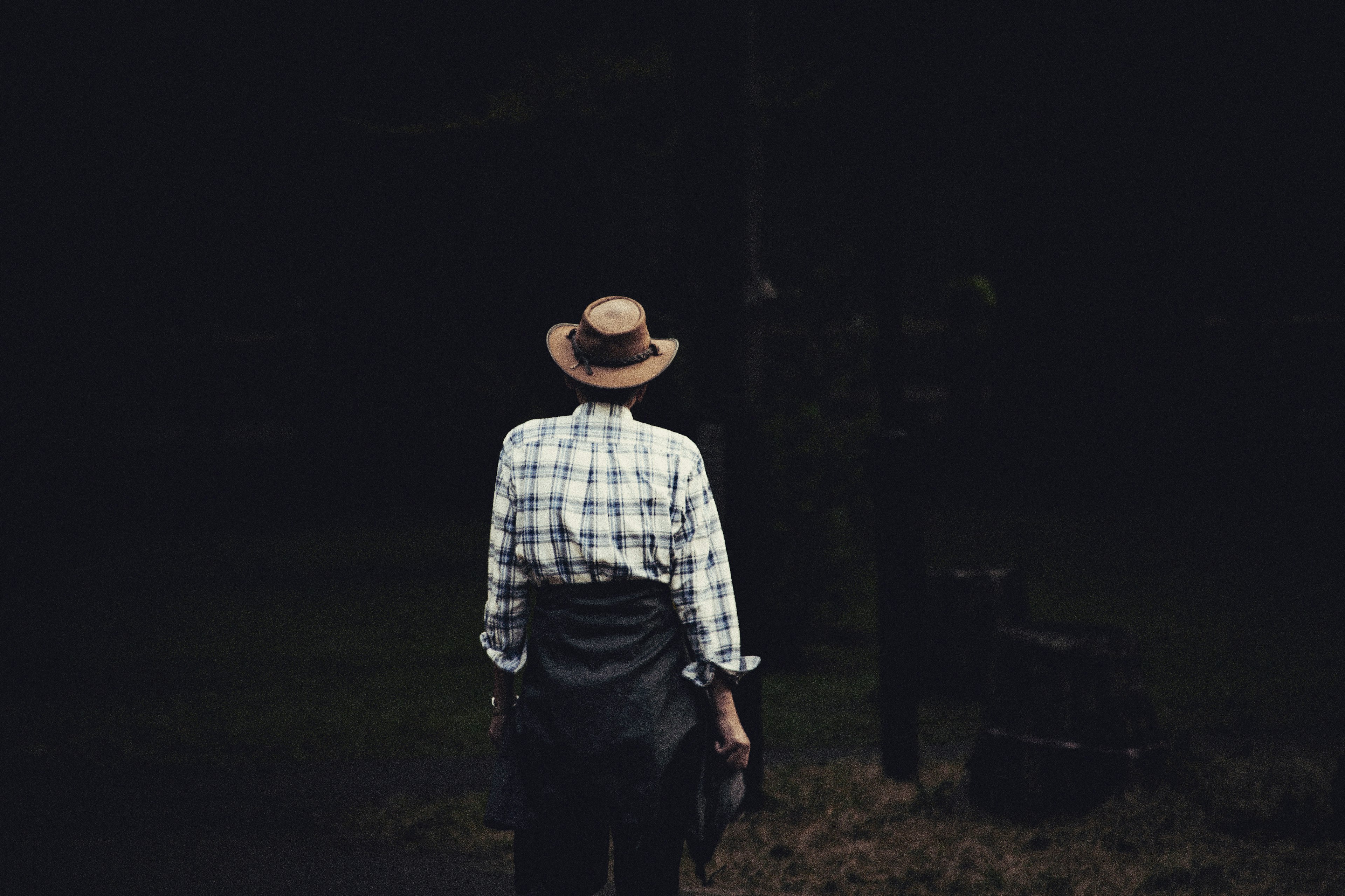 A man seen from behind wearing a hat and checkered shirt standing against a dark background