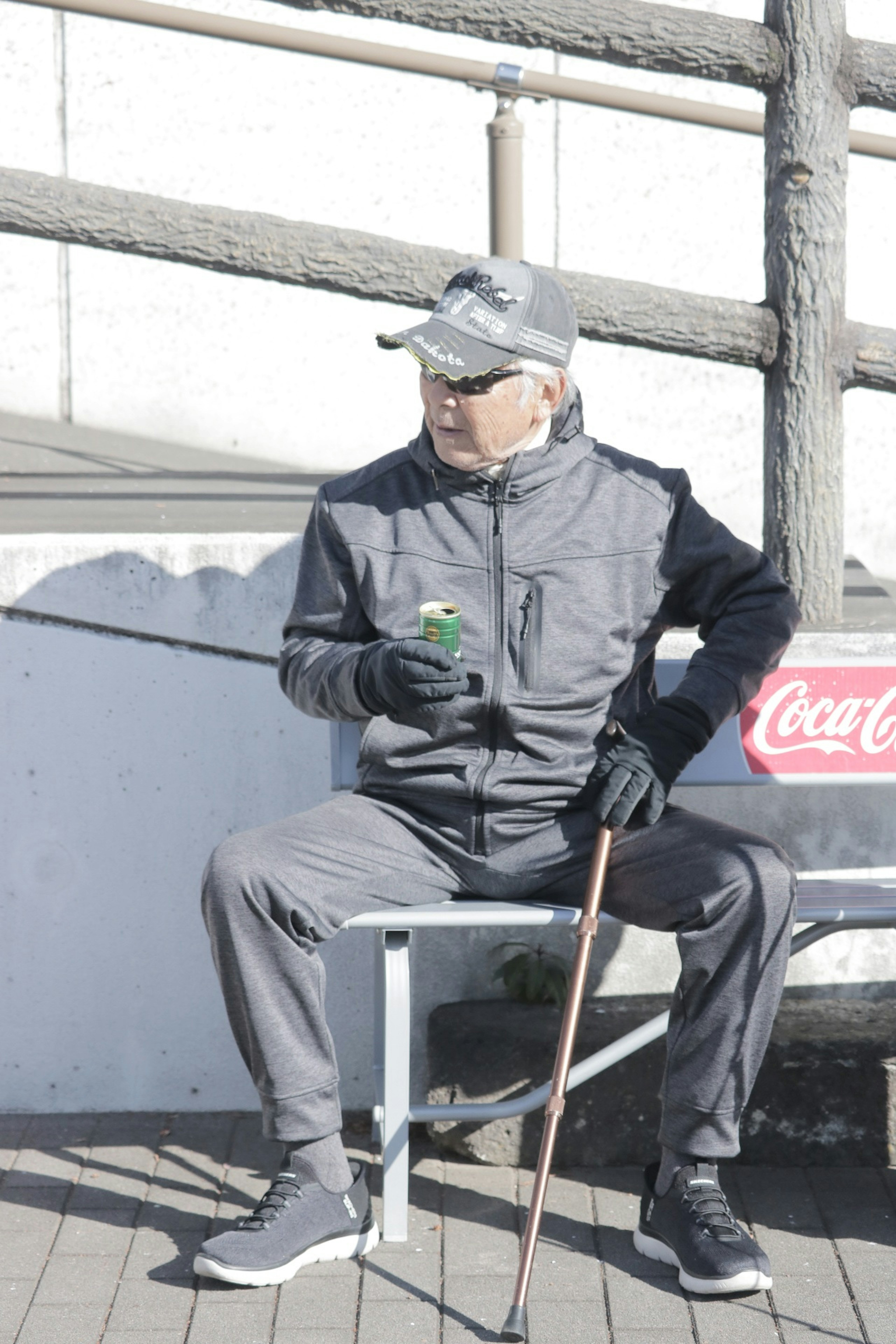 Seorang pria tua mengenakan jaket dan celana abu-abu duduk di depan papan Coca-Cola