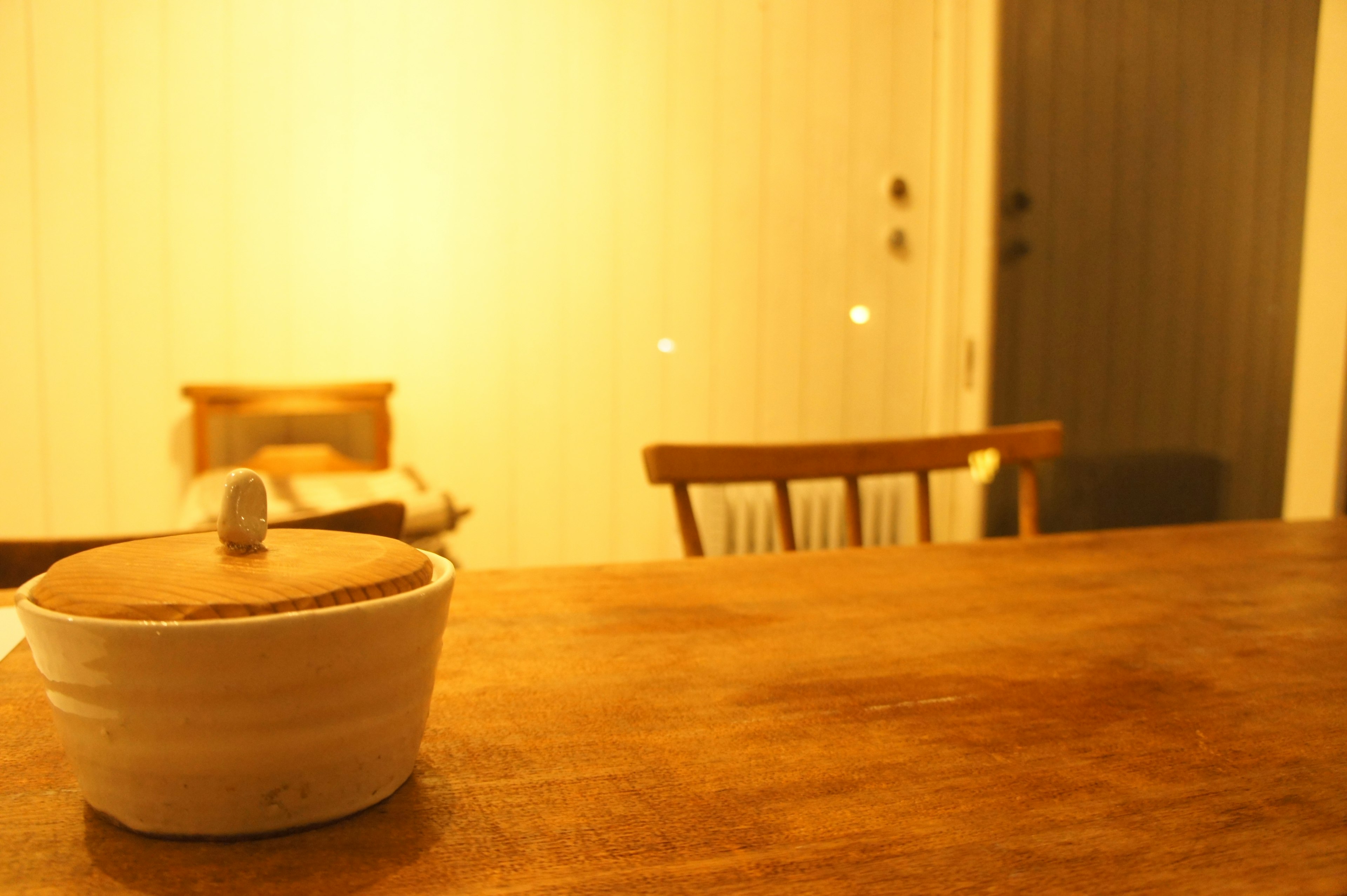 Warm indoor scene featuring a white ceramic container on a wooden table and a wooden chair in the background