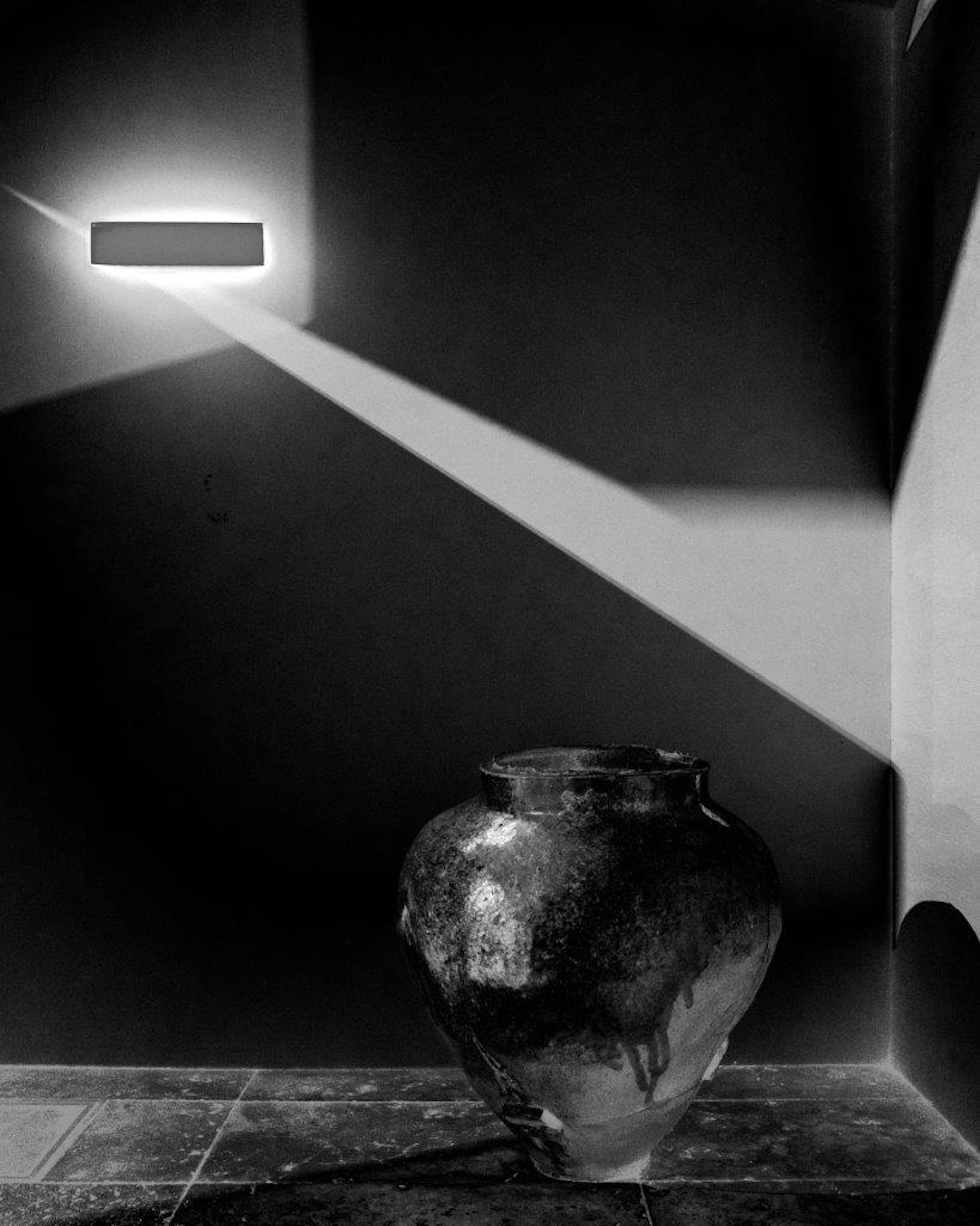 Black and white photo featuring a ceramic pot in the corner with a diagonal beam of light from the wall