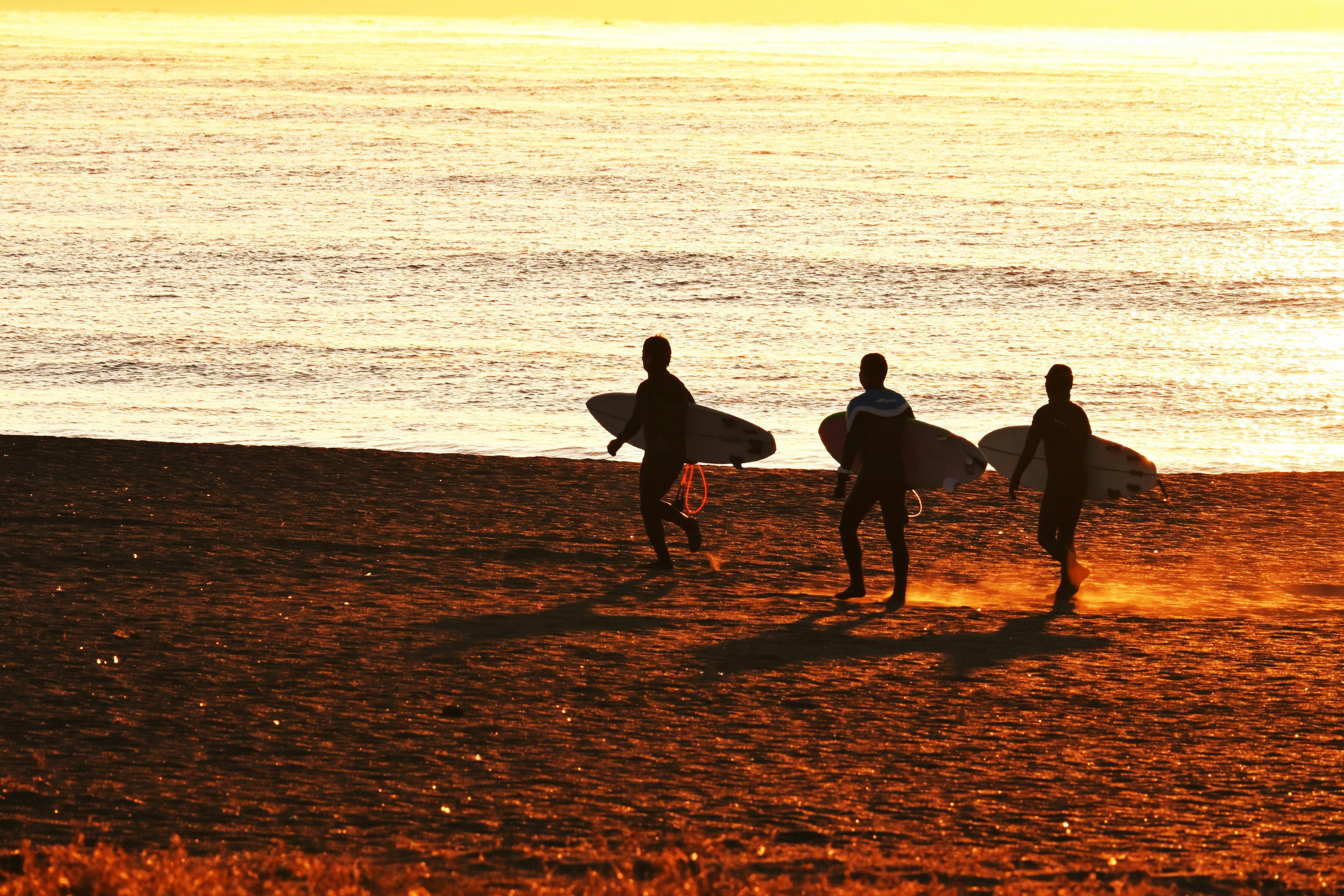 Three surfers carrying surfboards running towards the ocean at sunset