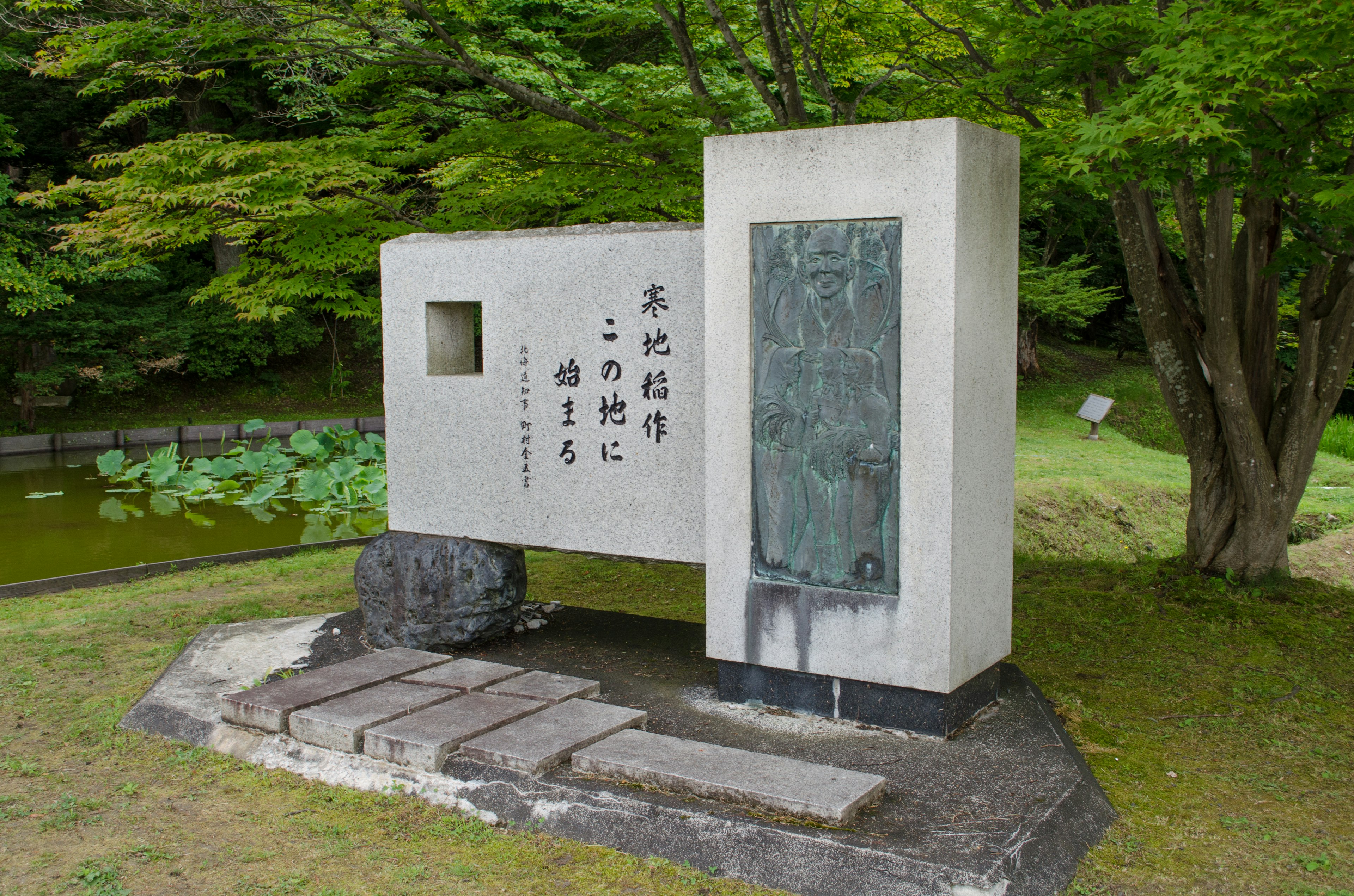 A stone monument in a lush green park featuring a sculpture and Japanese text