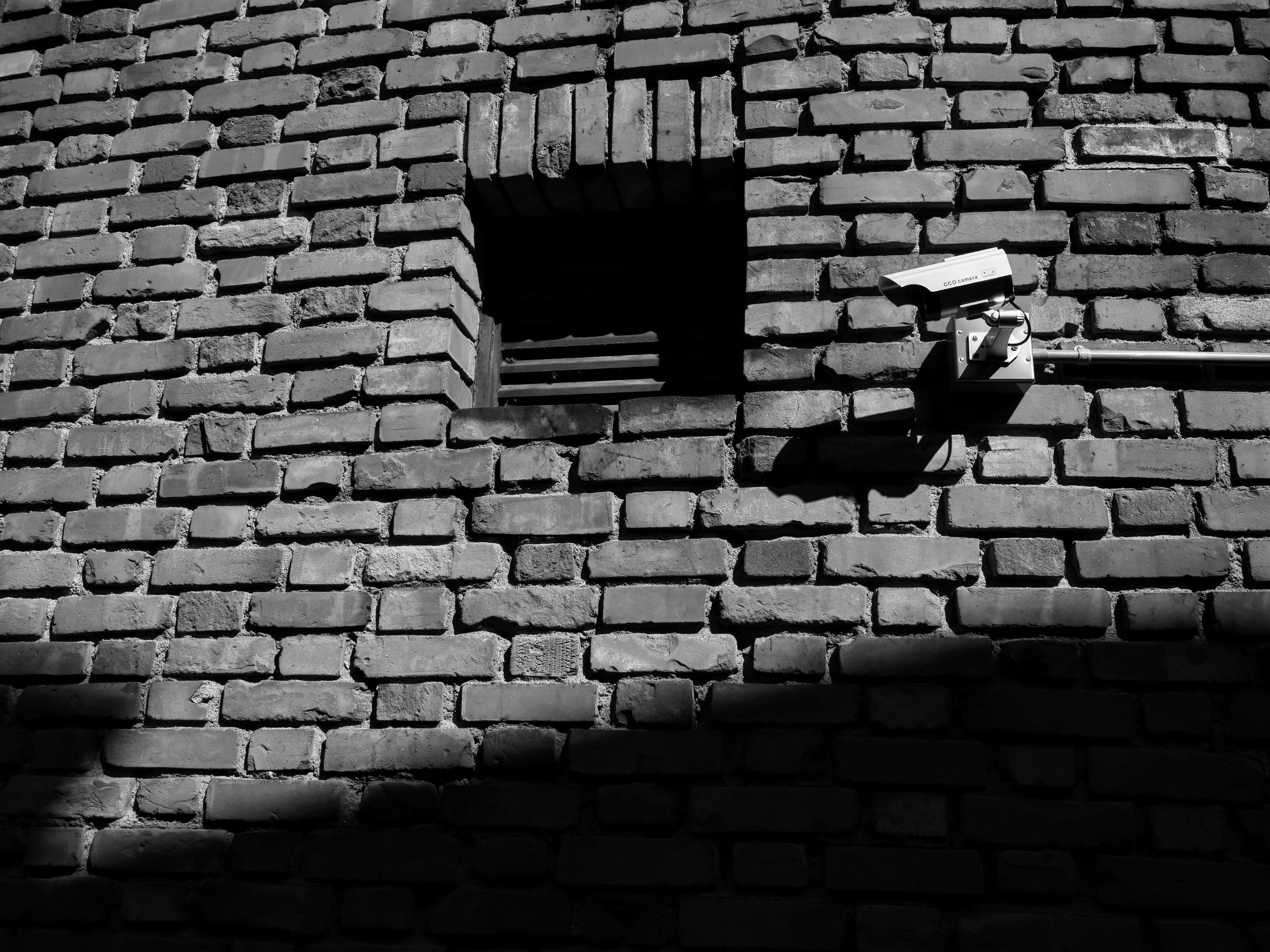 Pared de ladrillos en blanco y negro con una pequeña ventana y una cámara de seguridad