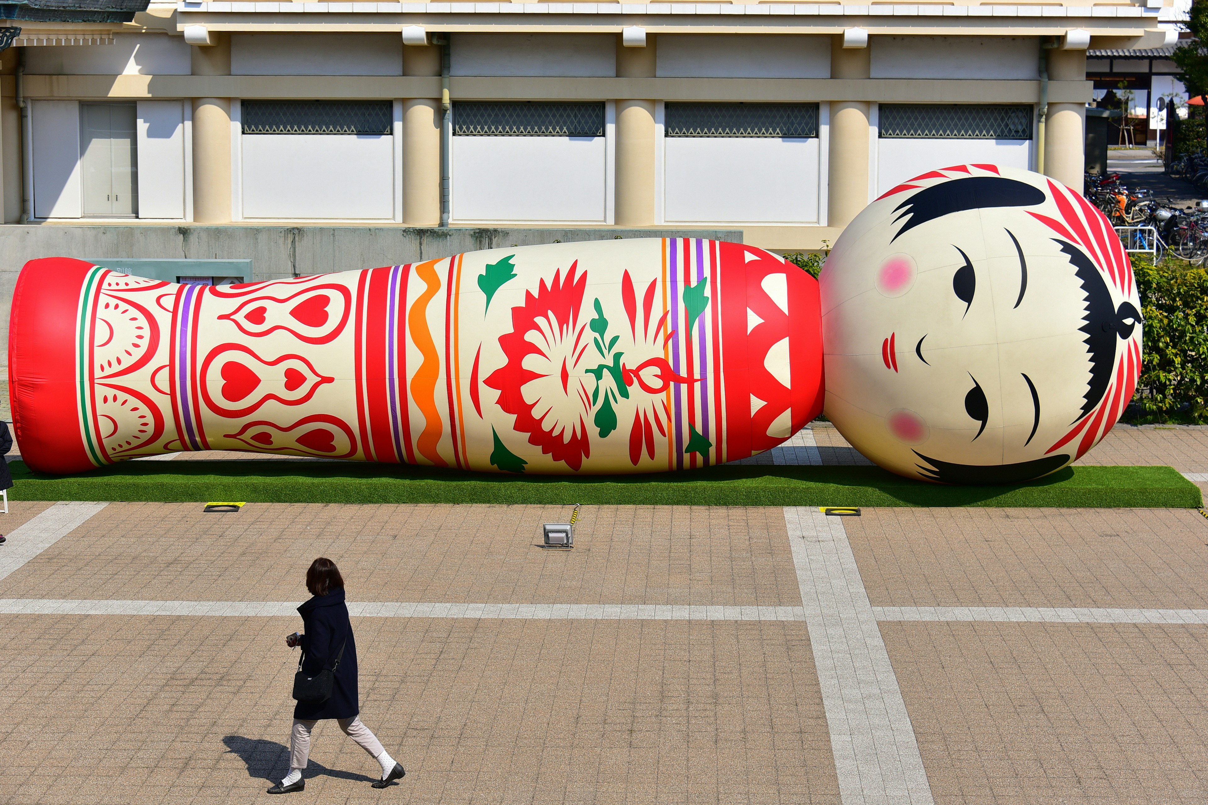 Una mujer caminando junto a una gran muñeca Kokeshi