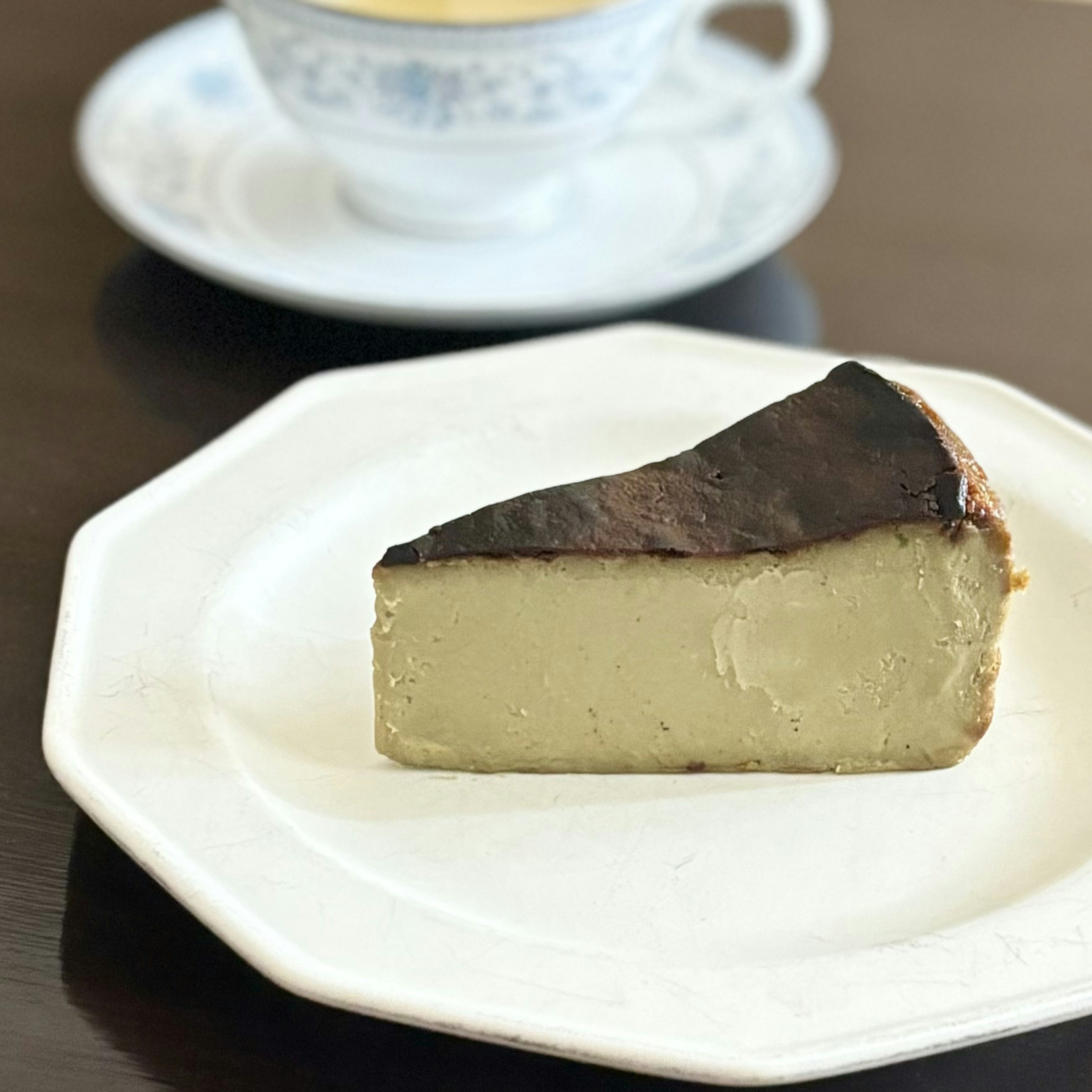 Ein Stück verbrannter Käsekuchen auf einem Teller mit einer Teetasse im Hintergrund