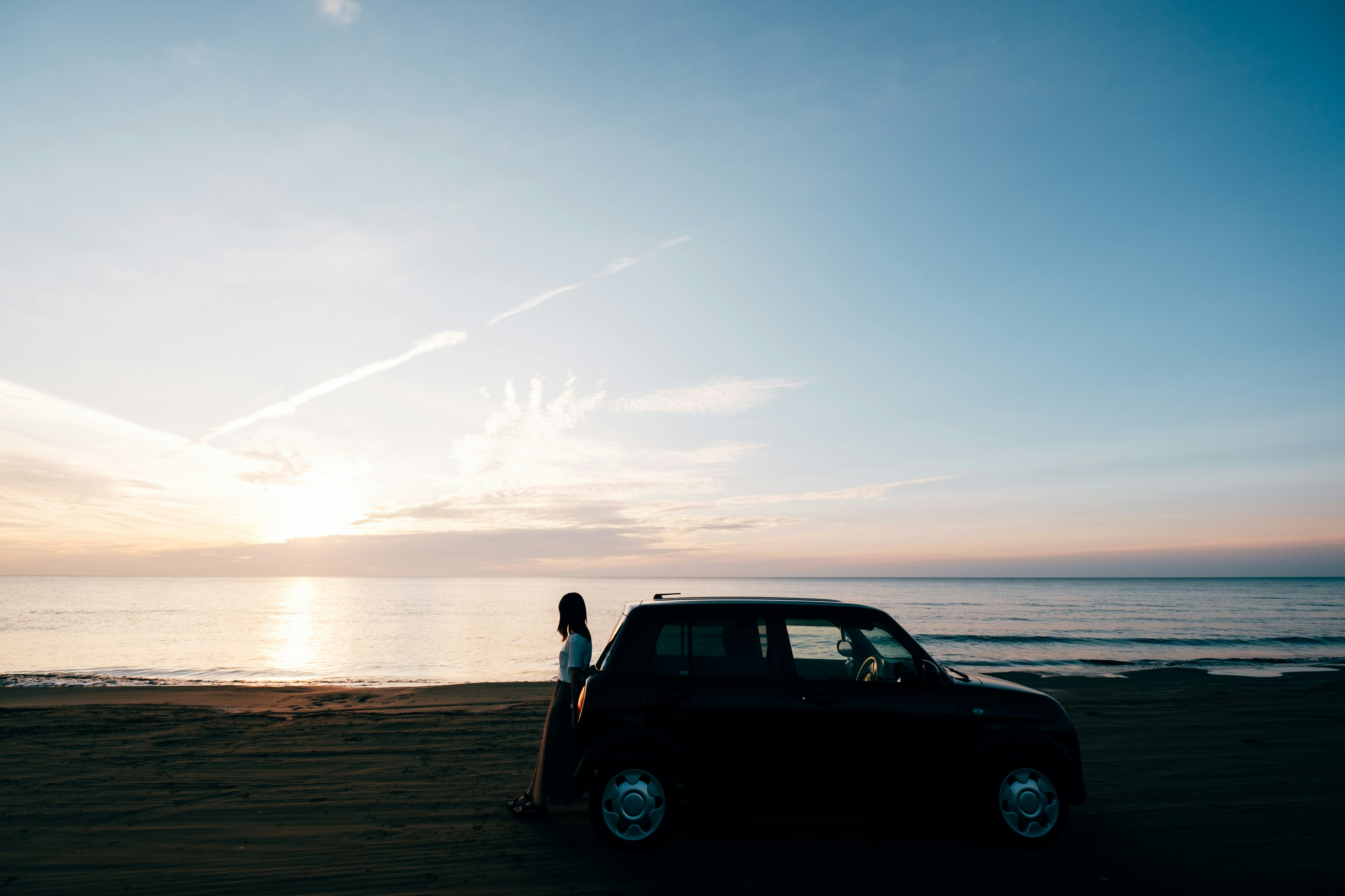 Une voiture noire avec une femme debout à côté d'elle au coucher du soleil sur la plage