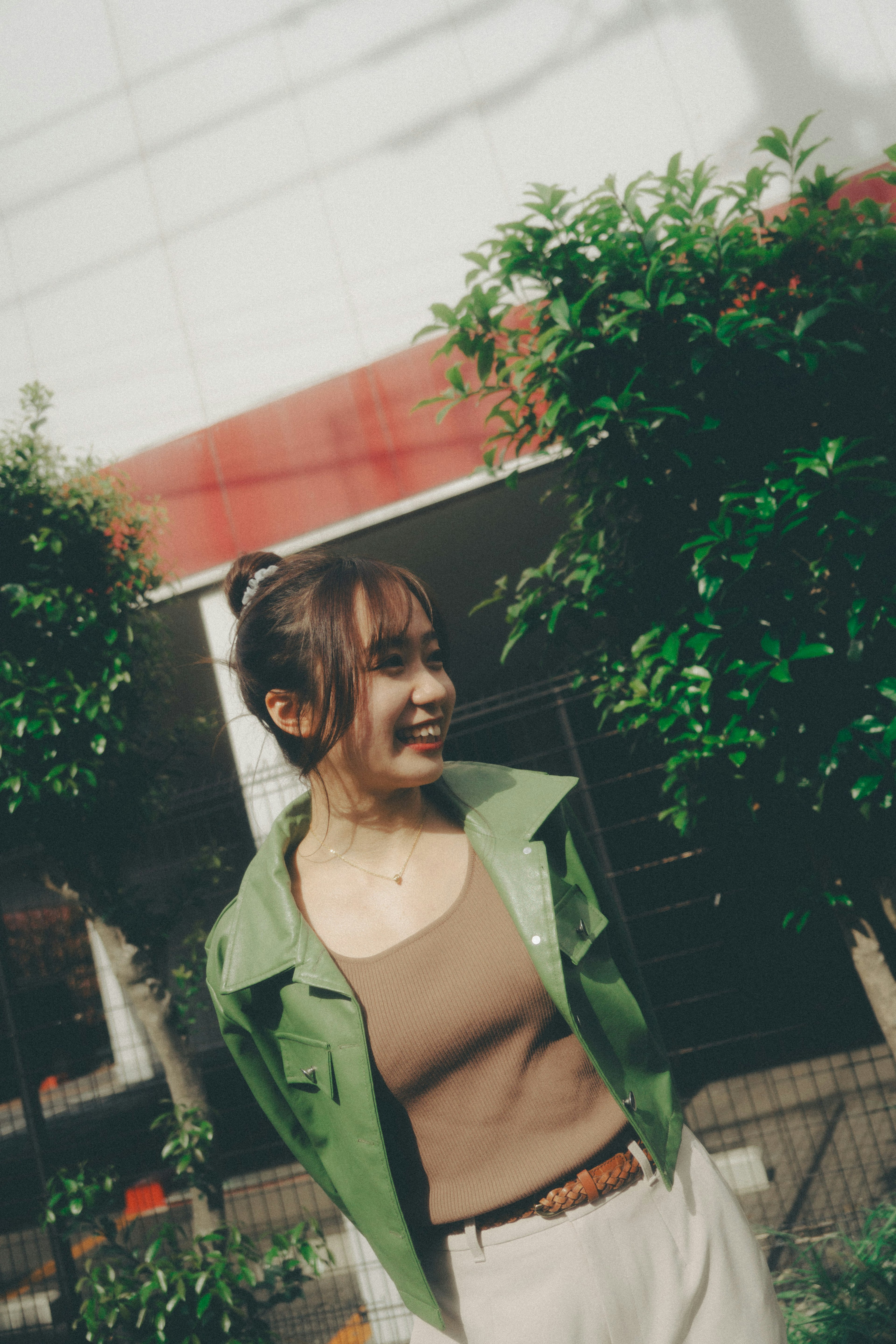 A smiling woman wearing a green jacket stands in front of green trees and a red building