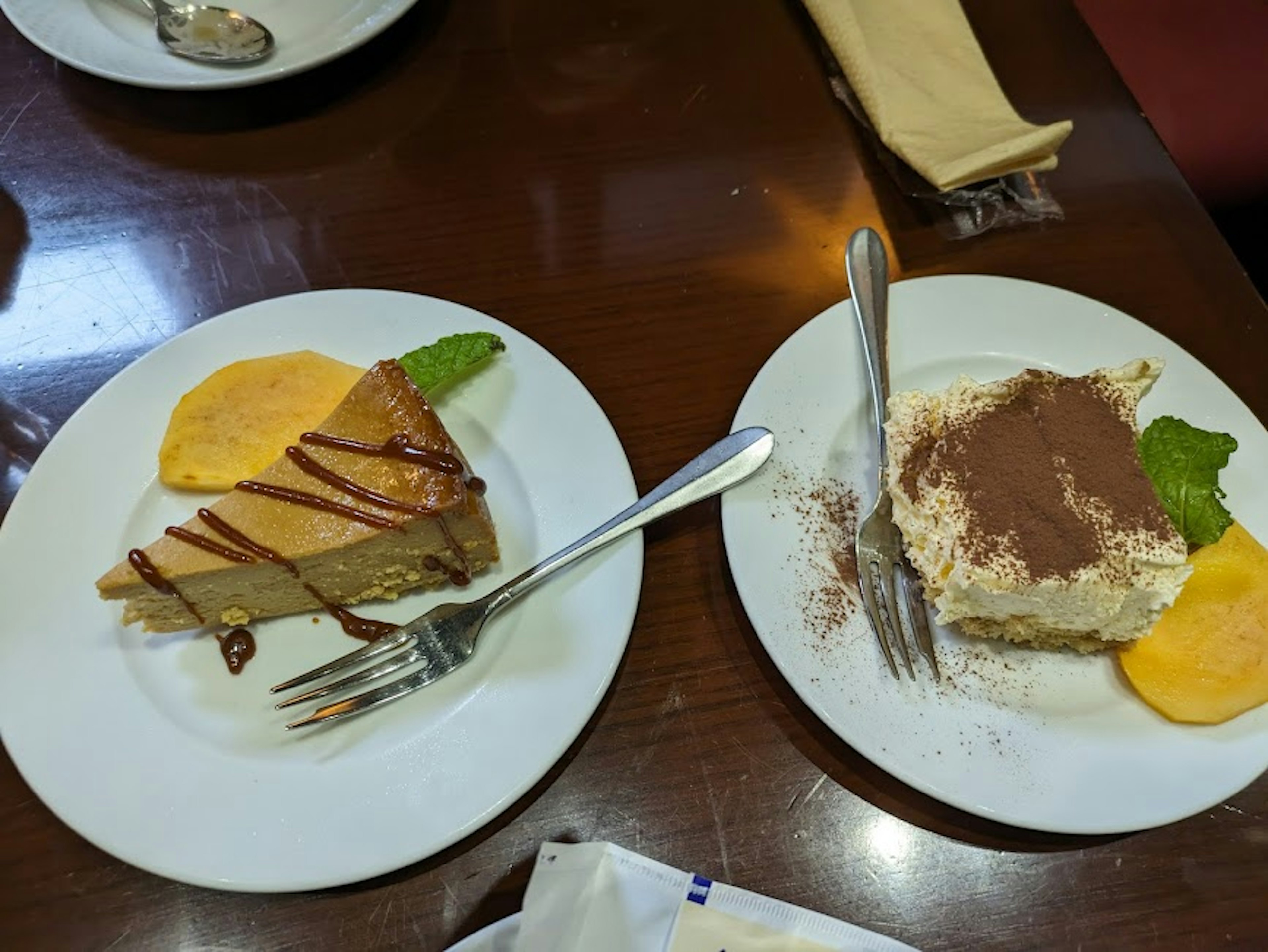 Dos platos de postre en una mesa Uno presenta un pastel cubierto de caramelo con una rodaja de naranja y hojas de menta El otro es tiramisú cubierto con cacao en polvo y hojas de menta