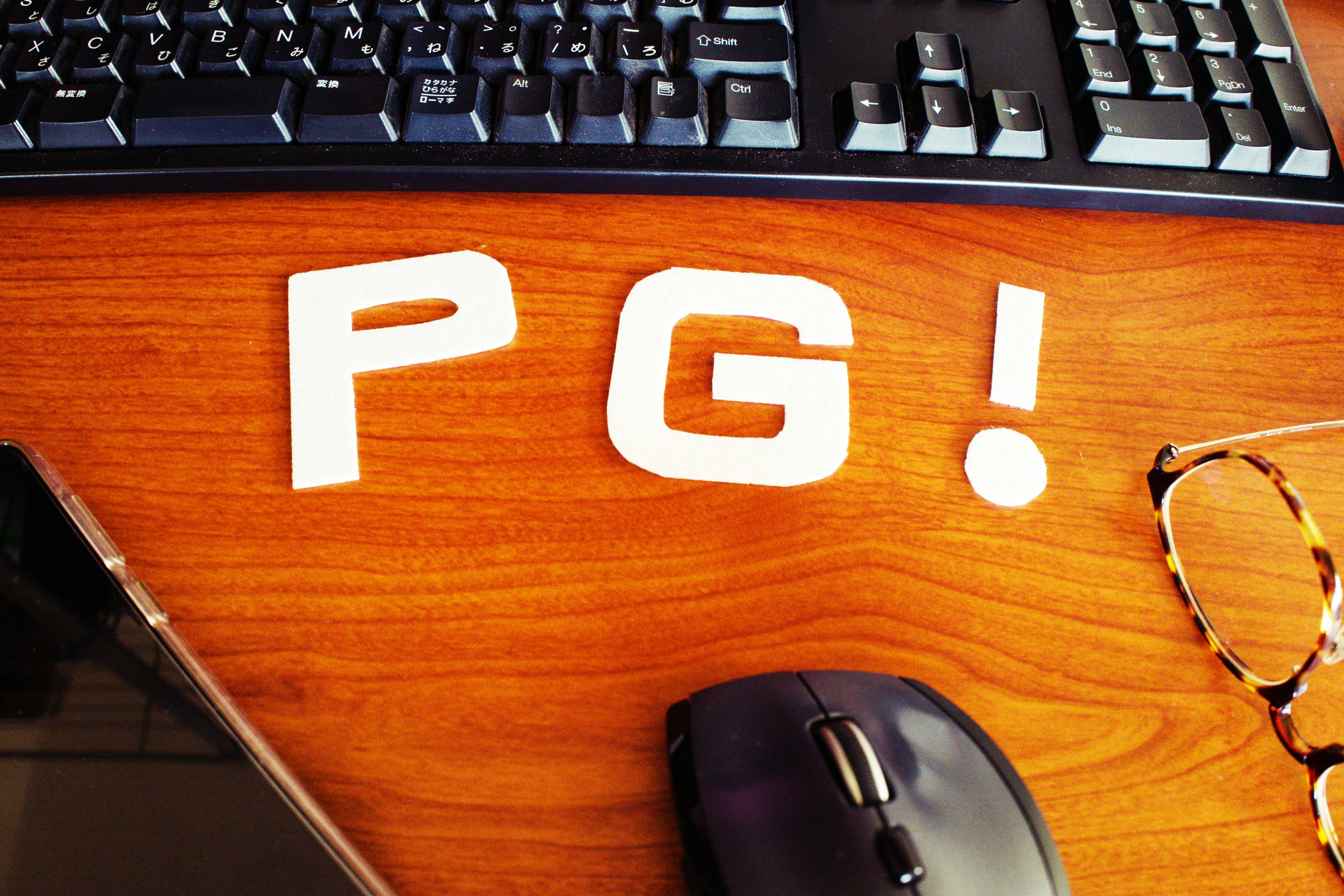 木製のデスクの上に置かれたキーボードとマウスの近くにある白い文字の「PG!」