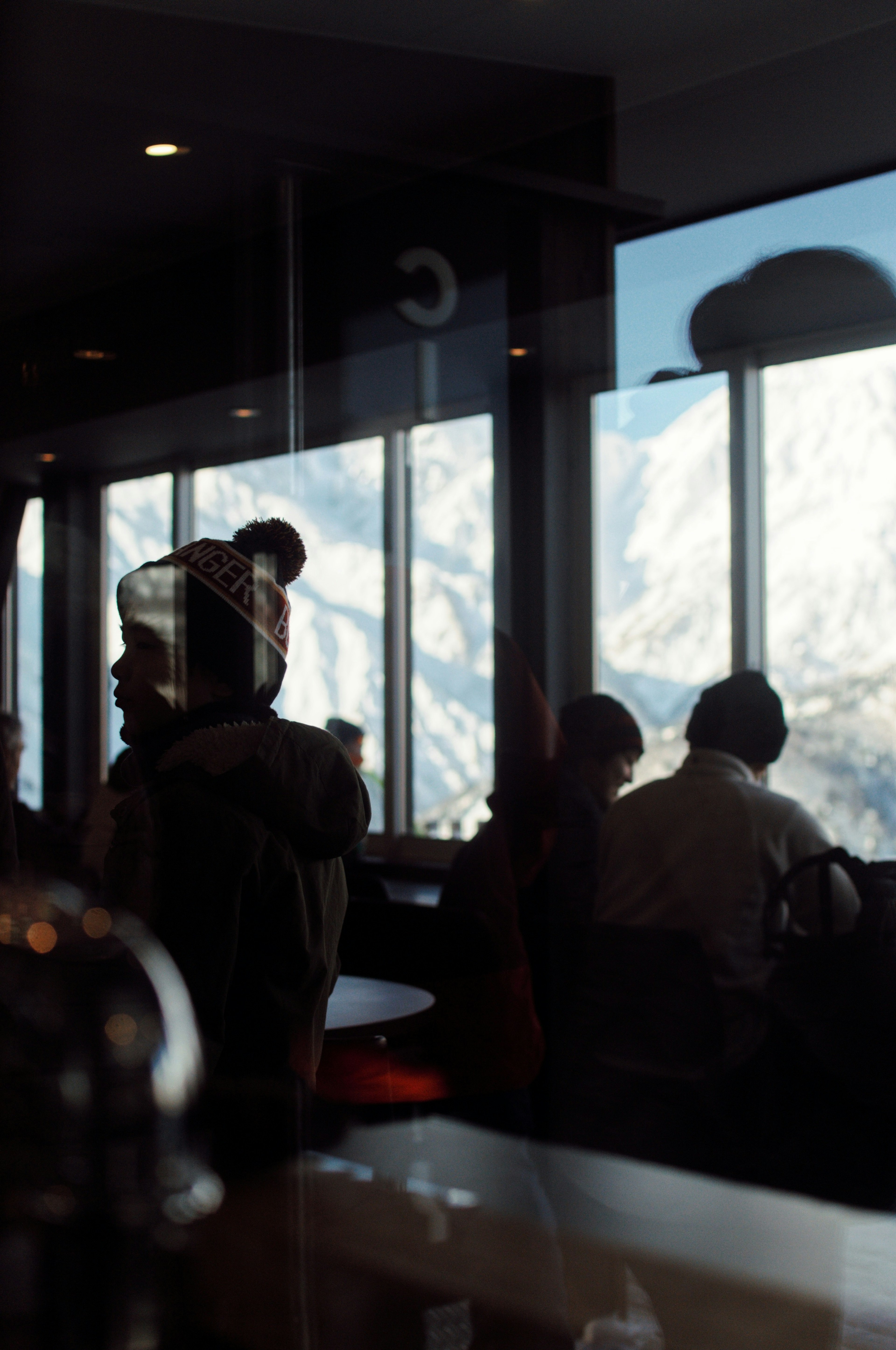 Innenansicht eines Cafés mit Menschen, die vor einer Kulisse von schneebedeckten Bergen stehen, die durch das Fenster sichtbar sind