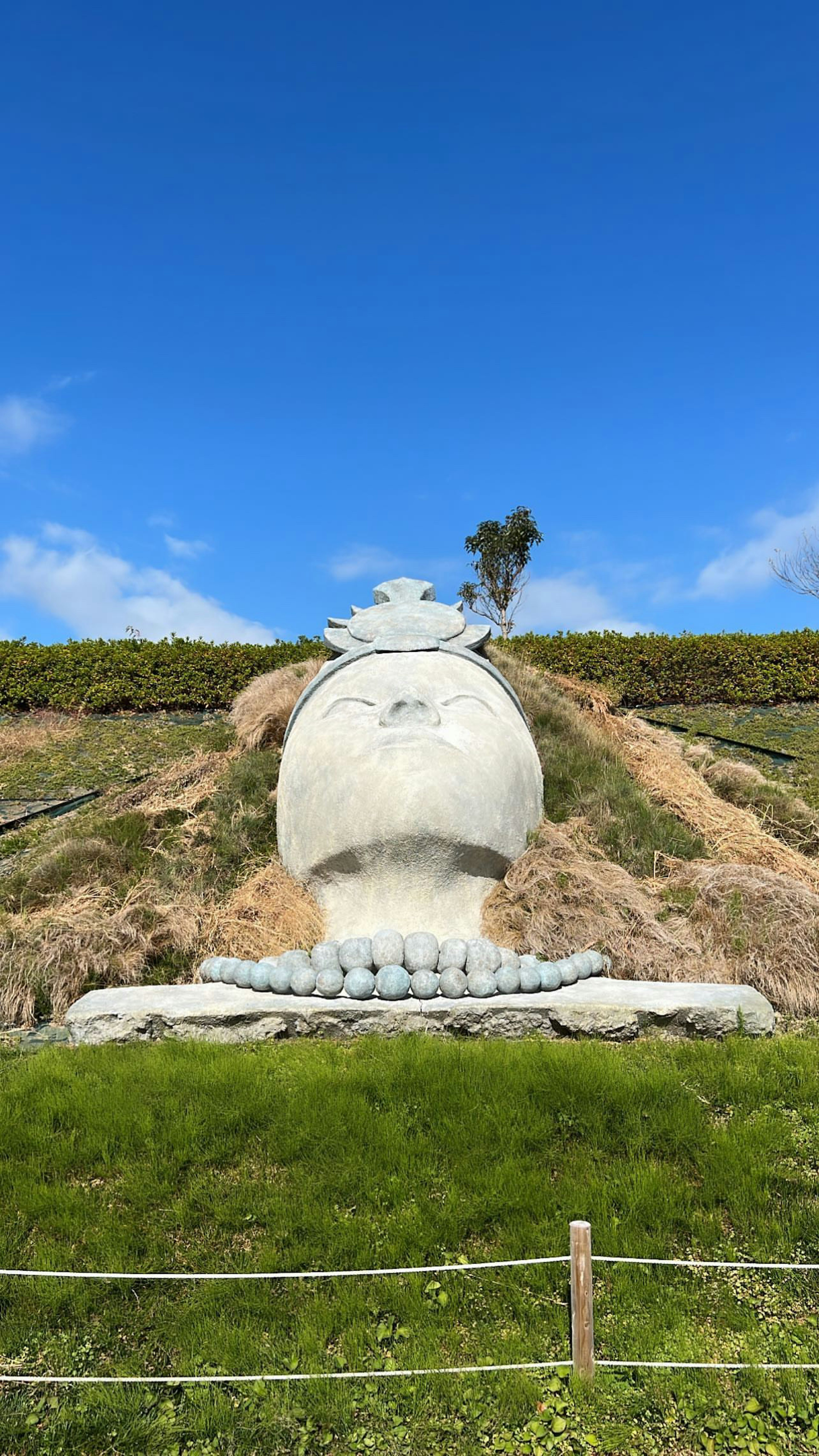 Large sculpture under a blue sky surrounded by green grass and hills with a tree in the distance