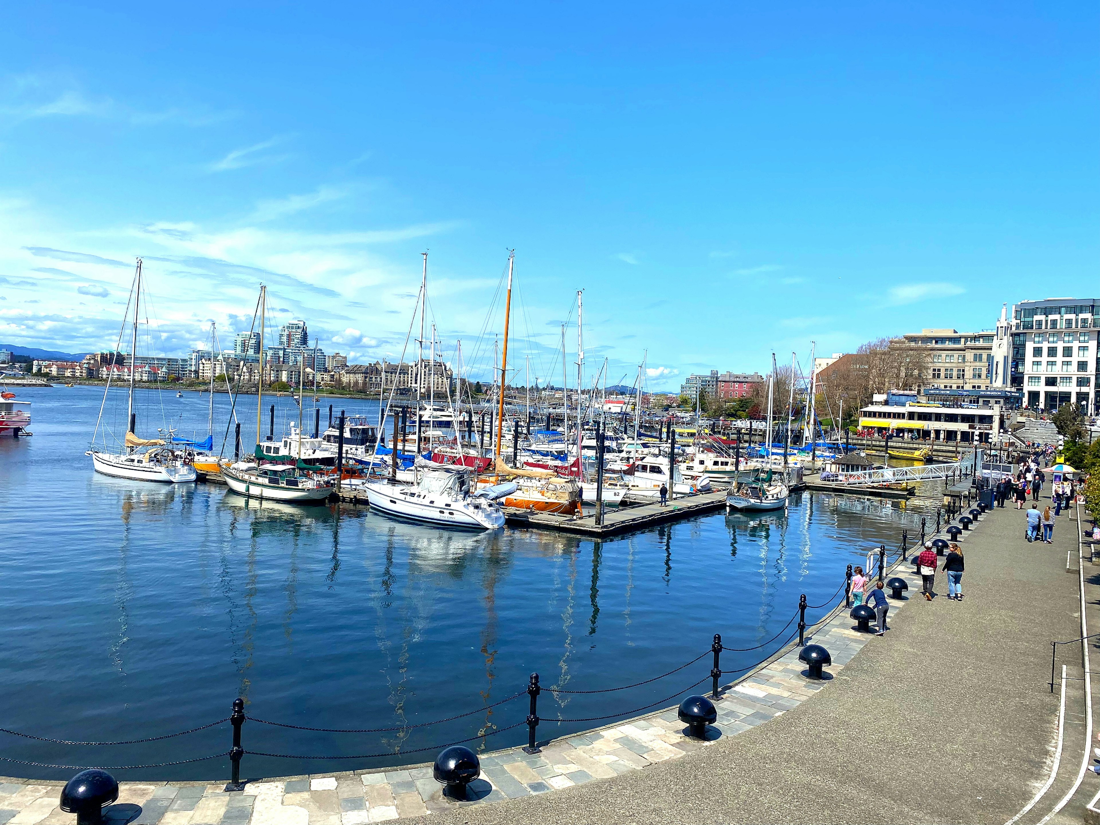 Vista del puerto con barcos amarrados y personas caminando por la costa bajo un cielo azul