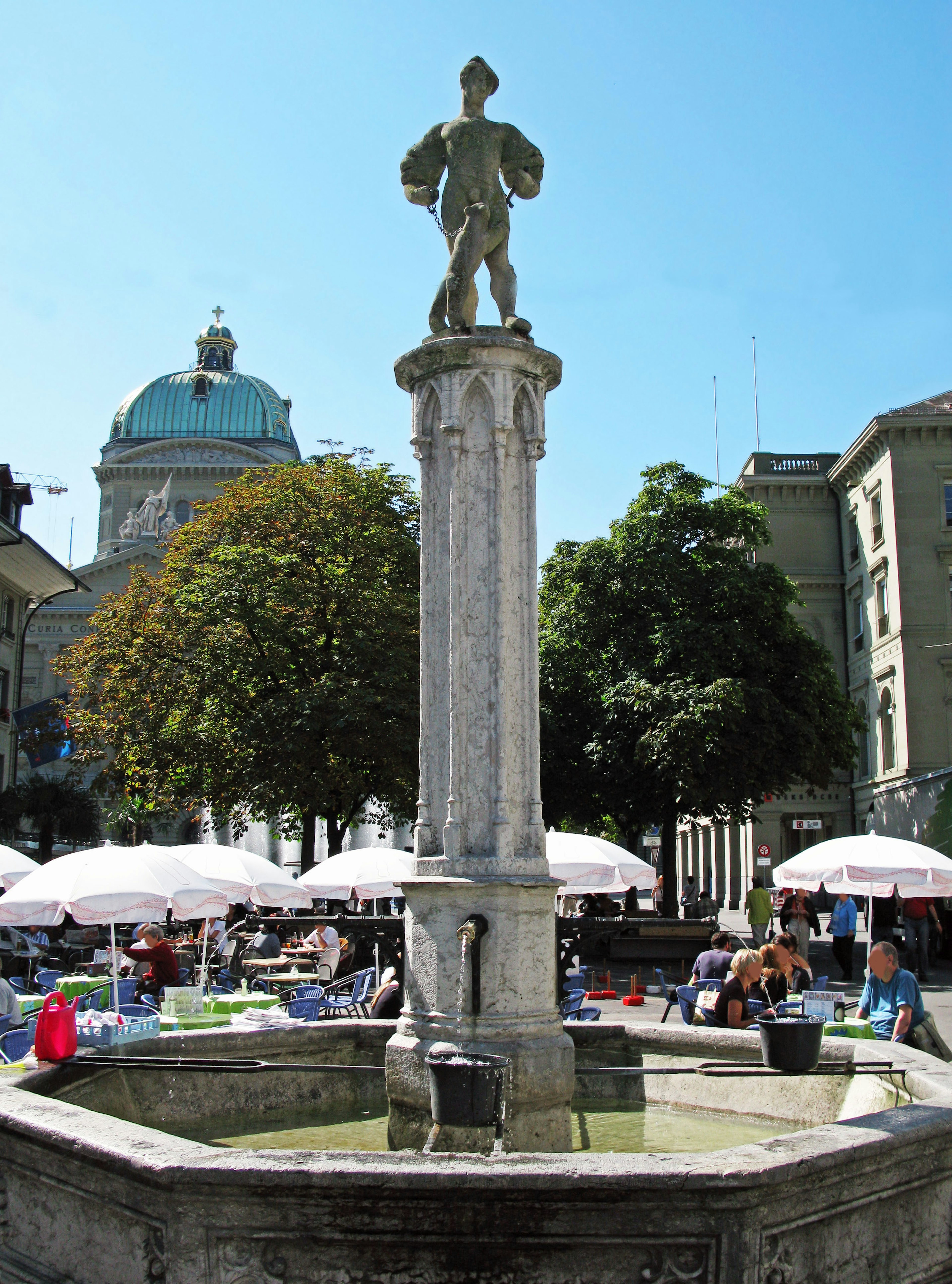 Fontaine avec une statue sous un ciel bleu dégagé entourée de personnes dans une place animée