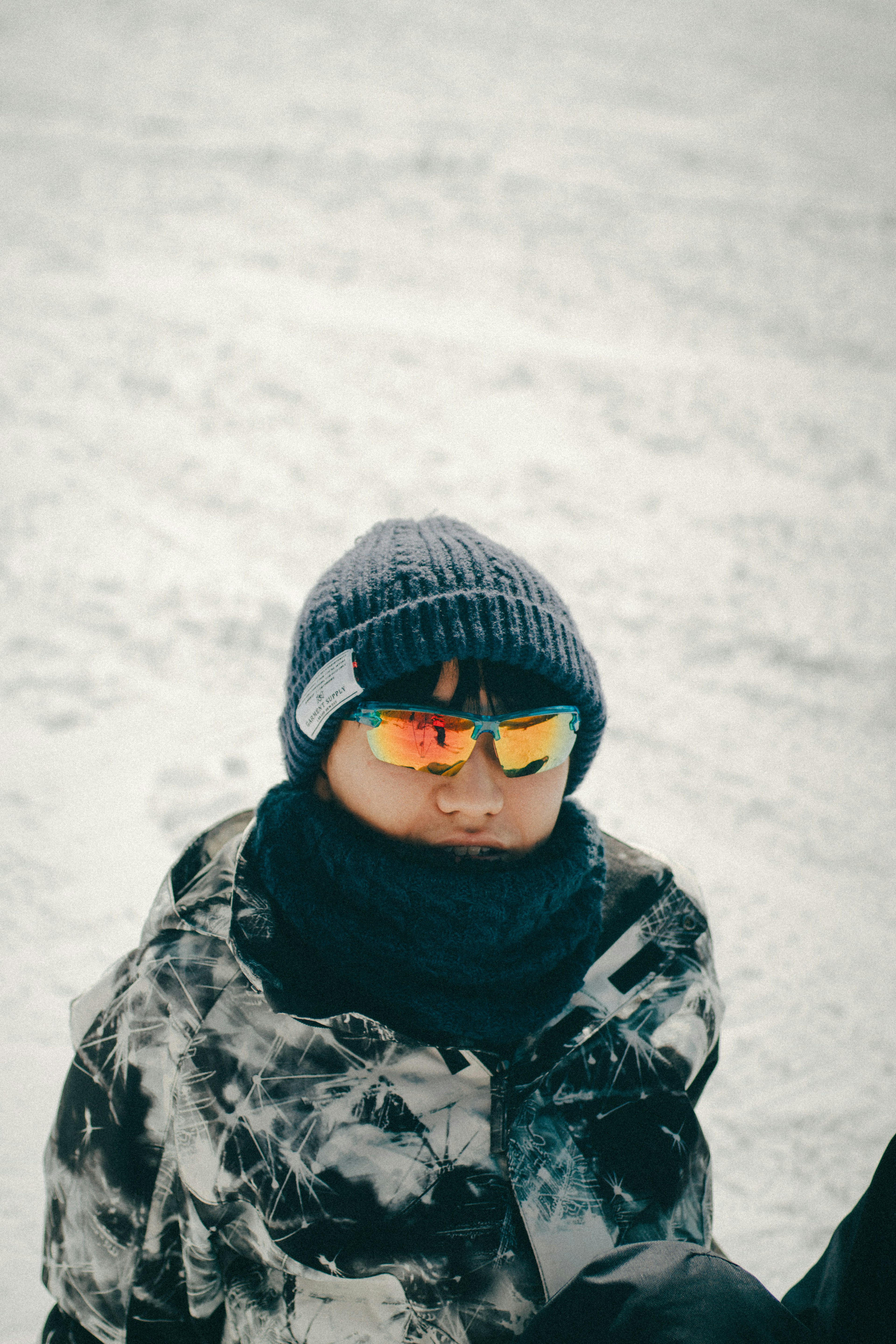 A boy sitting in the snow wearing a knitted hat and sunglasses