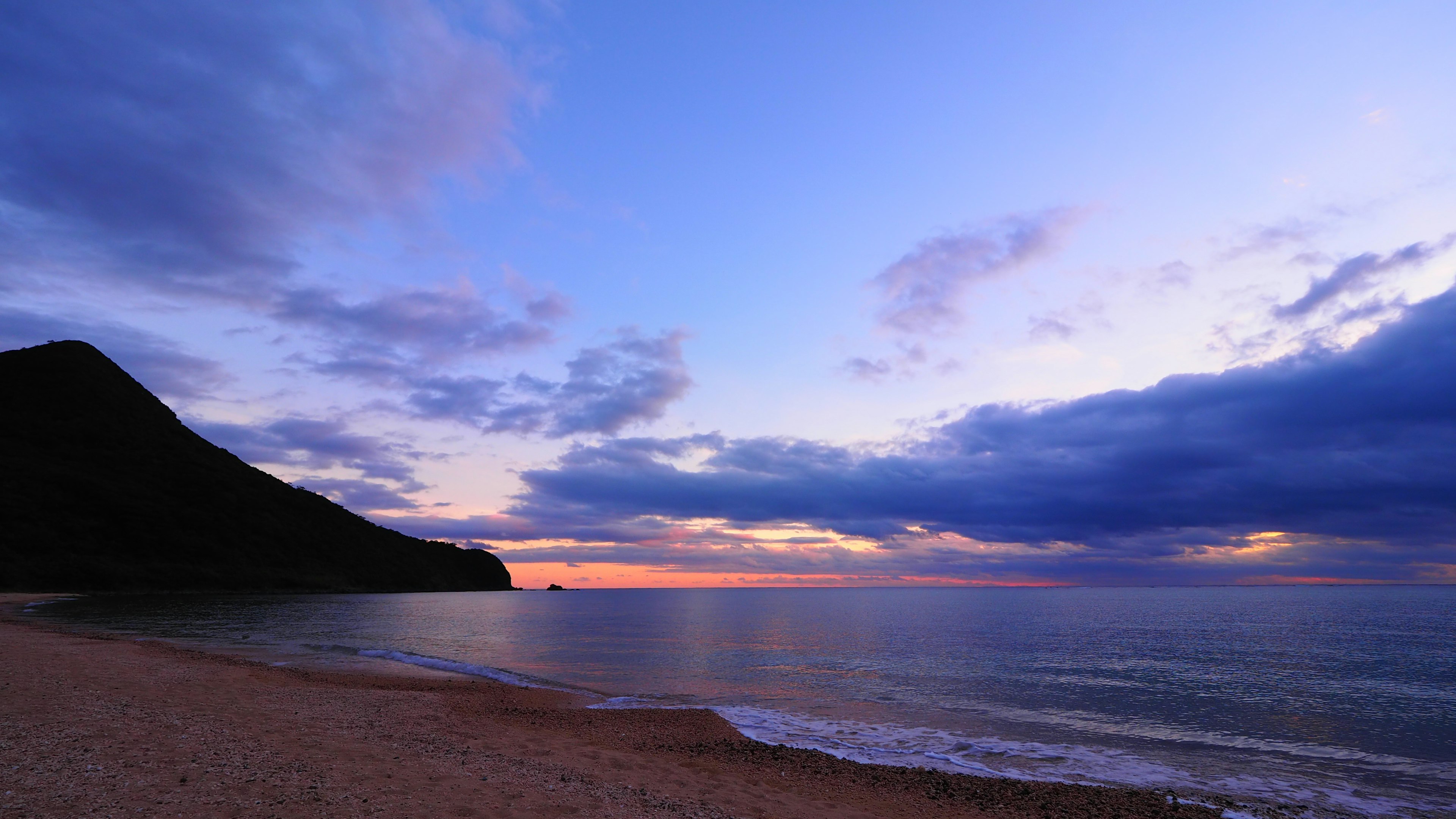 Pemandangan pantai saat senja dengan siluet gunung dan laut tenang