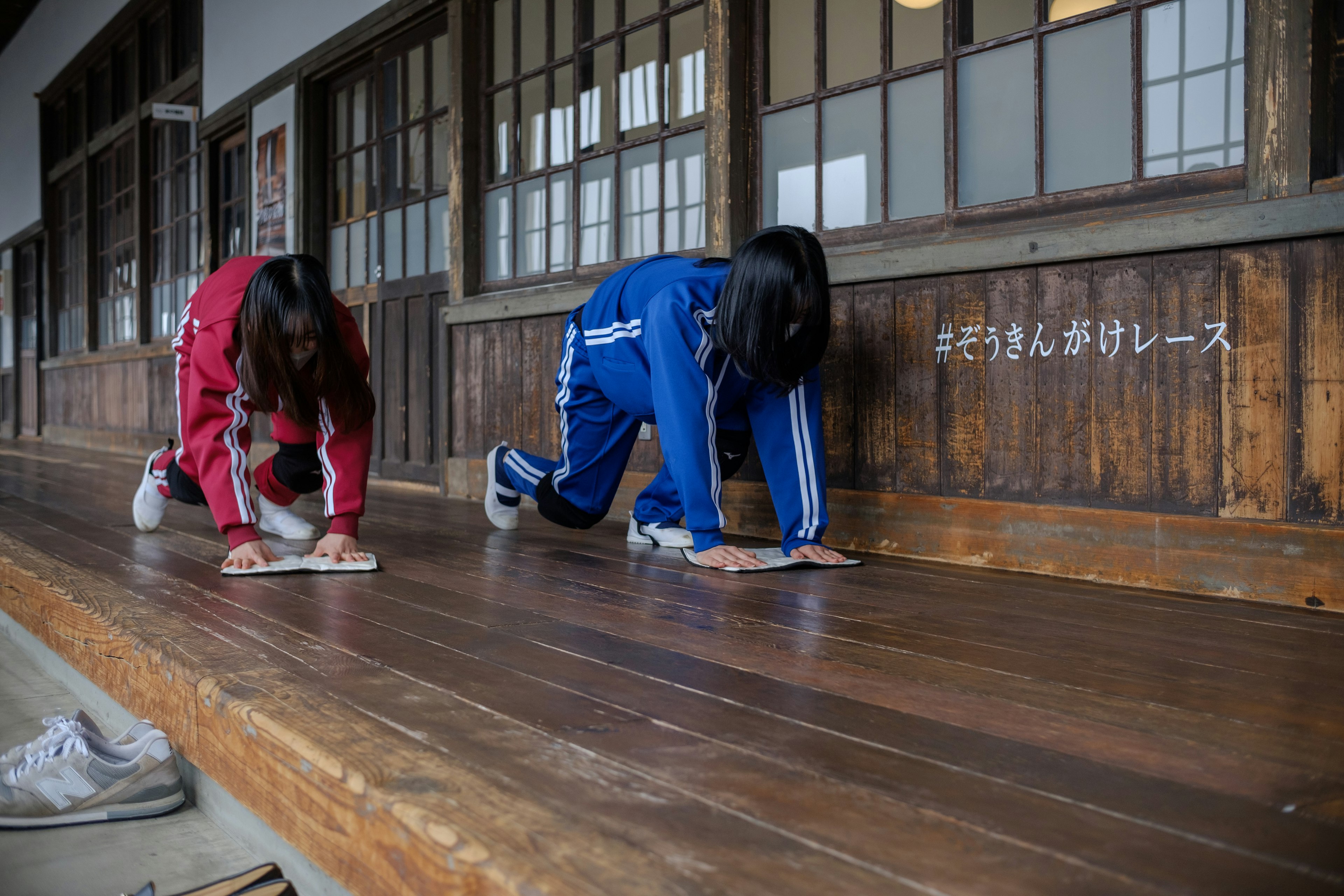 Dos estudiantes limpiando un pasillo escolar de madera con chándales rojo y azul