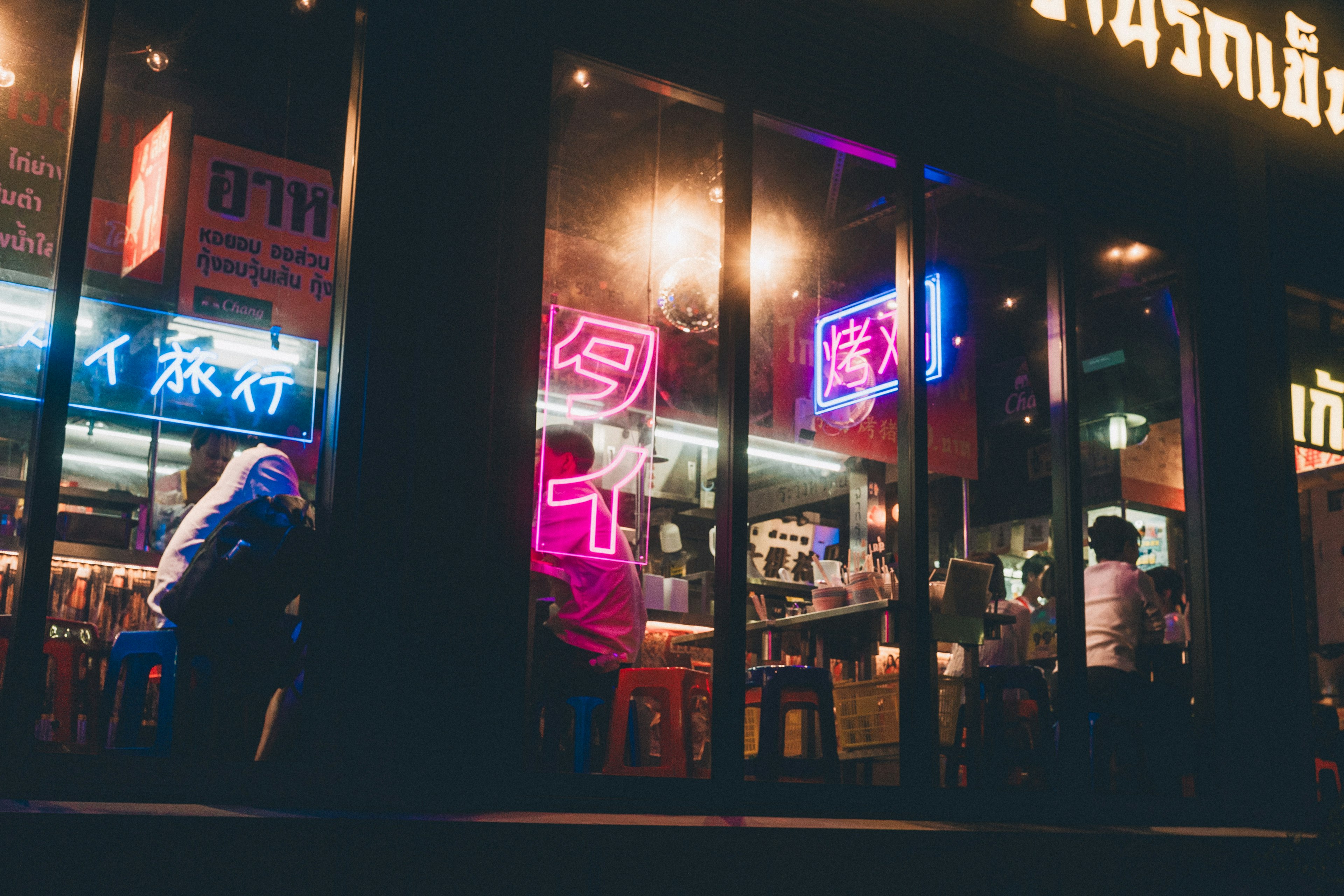 Exterior of a vibrant restaurant at night with glowing neon signs and people seated inside