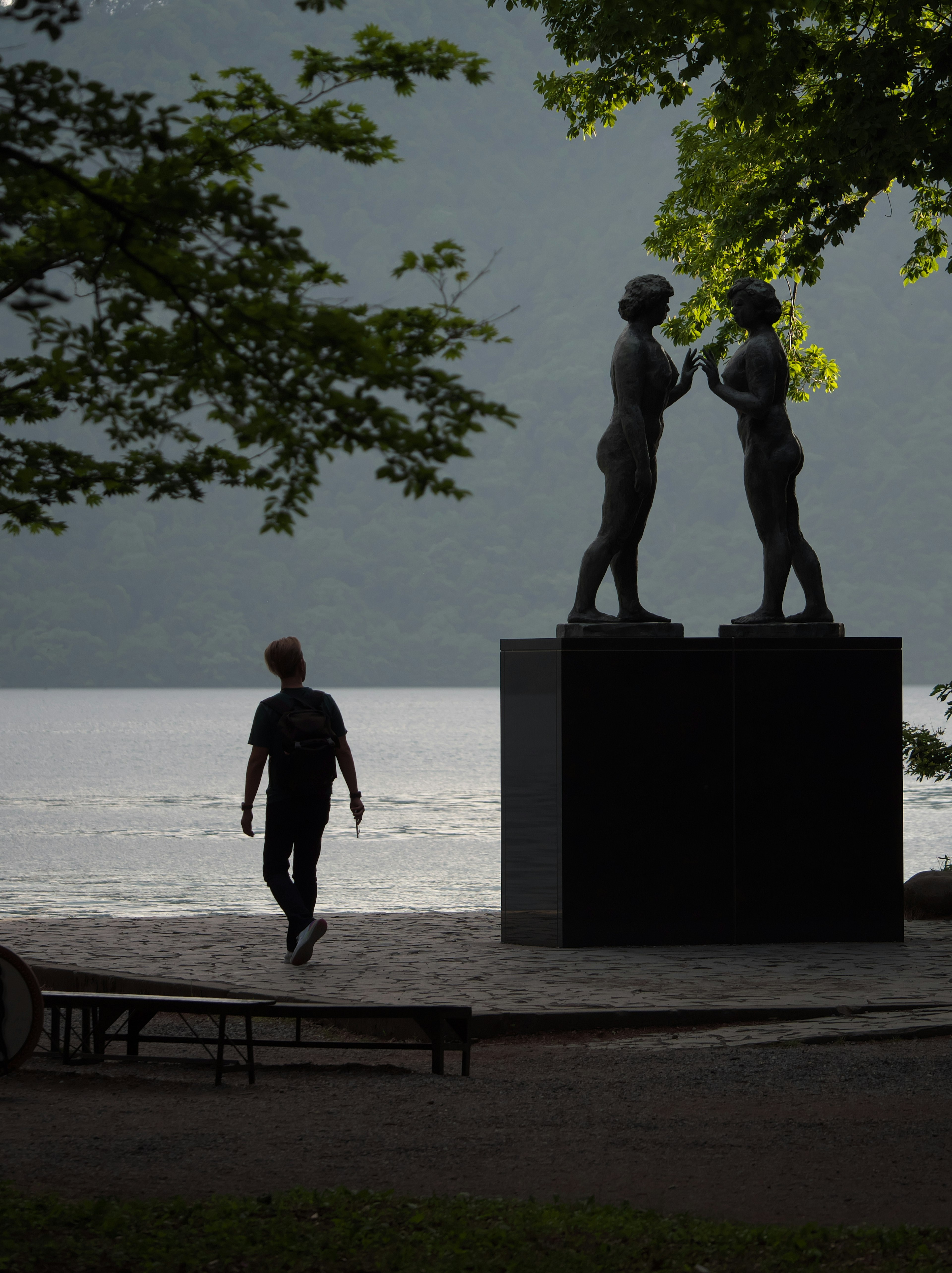 A person walking past two sculptures by a calm lake